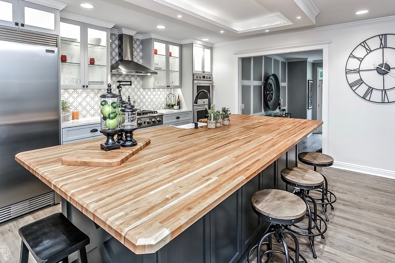Modern kitchen with a large wooden island, stainless steel appliances, white cabinetry, a decorative vent hood, and a large wall clock.