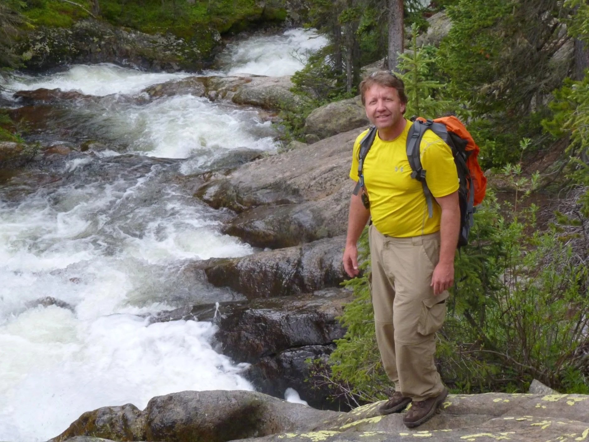 A man wearing a yellow shirt and beige pants standing on rocks near a rushing river in a forested area, carrying an orange backpack.