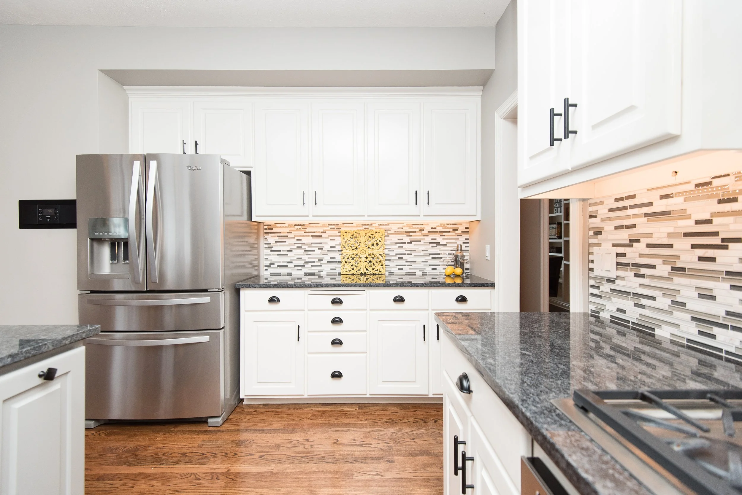 Modern kitchen with white cabinets, stainless steel refrigerator, black hardware, granite countertops, colorful backsplash, and hardwood floors.