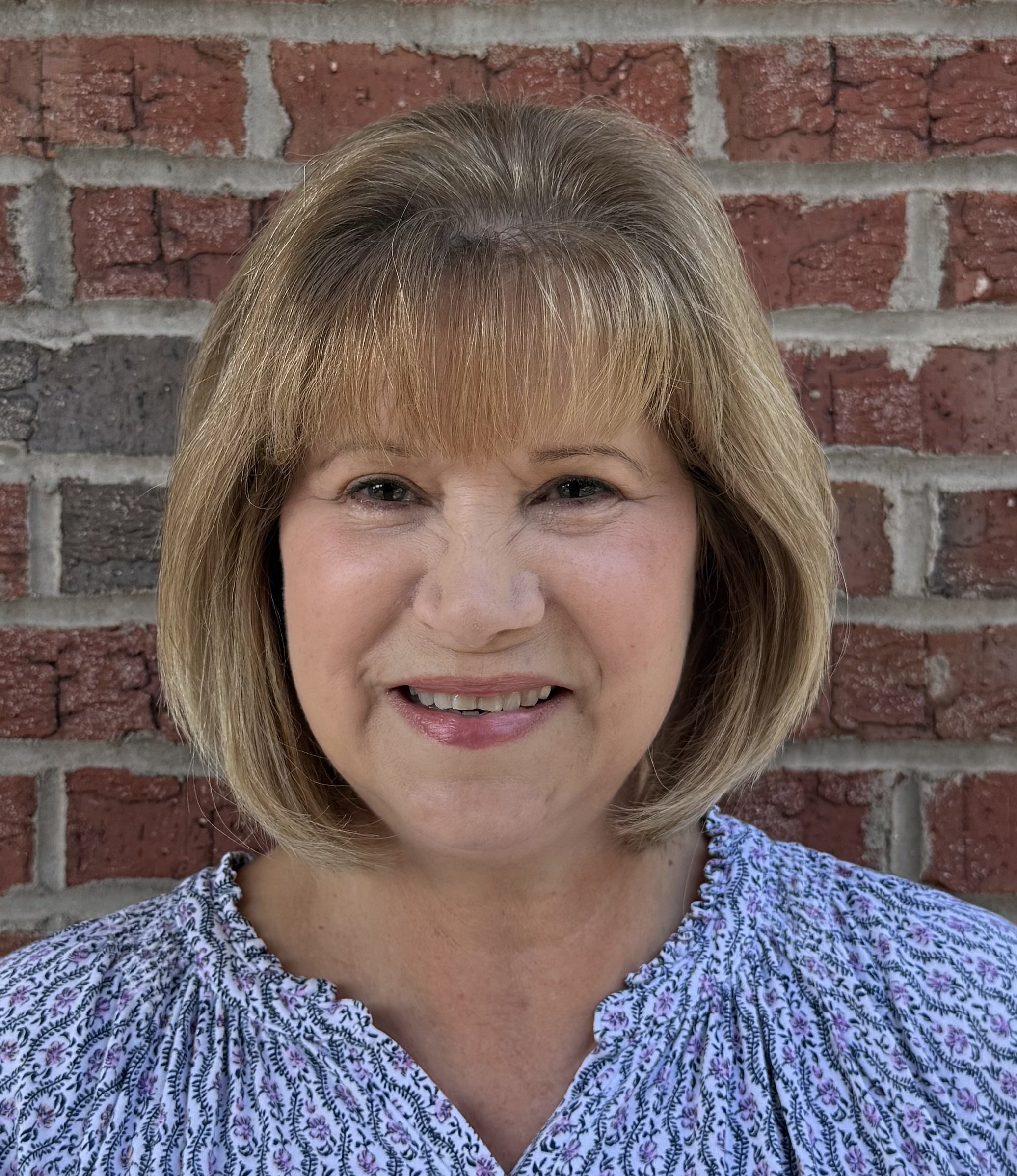 Close-up of a woman with light brown hair and a smile, wearing a patterned blouse, standing in front of a brick wall.