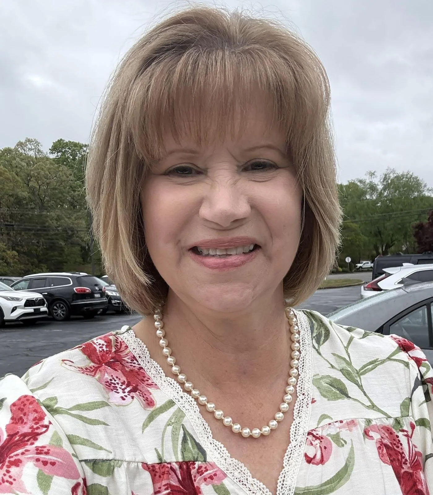 A smiling woman with short, light brown hair wearing a floral dress and pearl necklace. She is outdoors in a parking lot with cars and green trees in the background under cloudy skies.