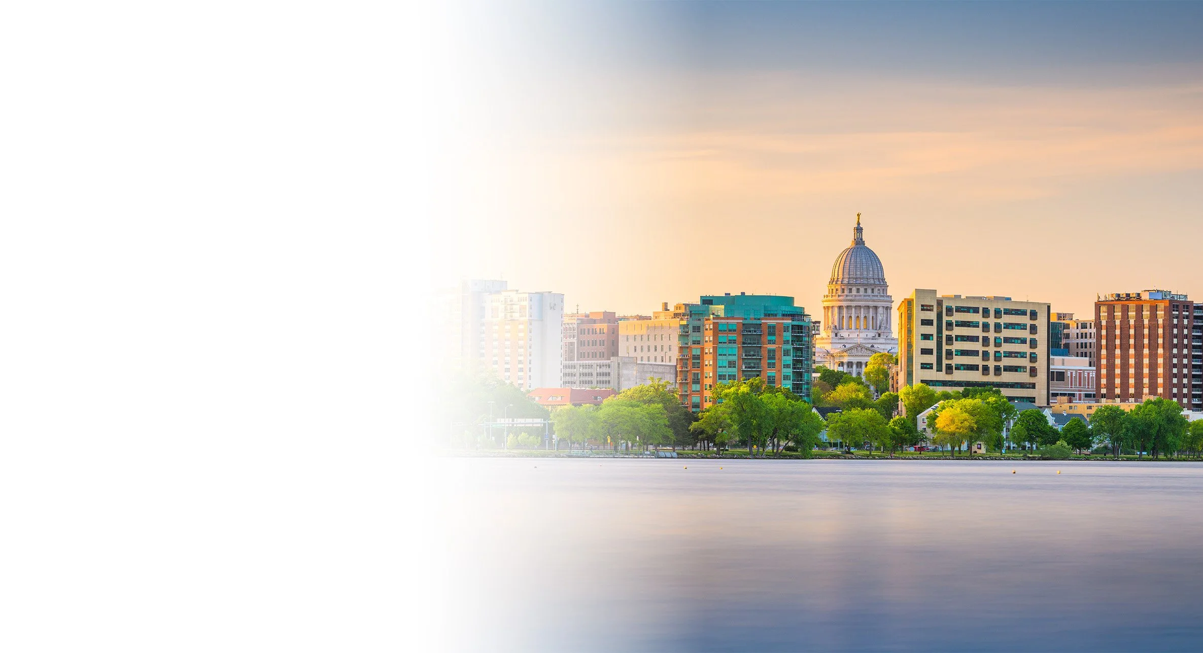 City skyline with a historic dome building, modern high-rises, and lush green trees along a waterfront during sunset.