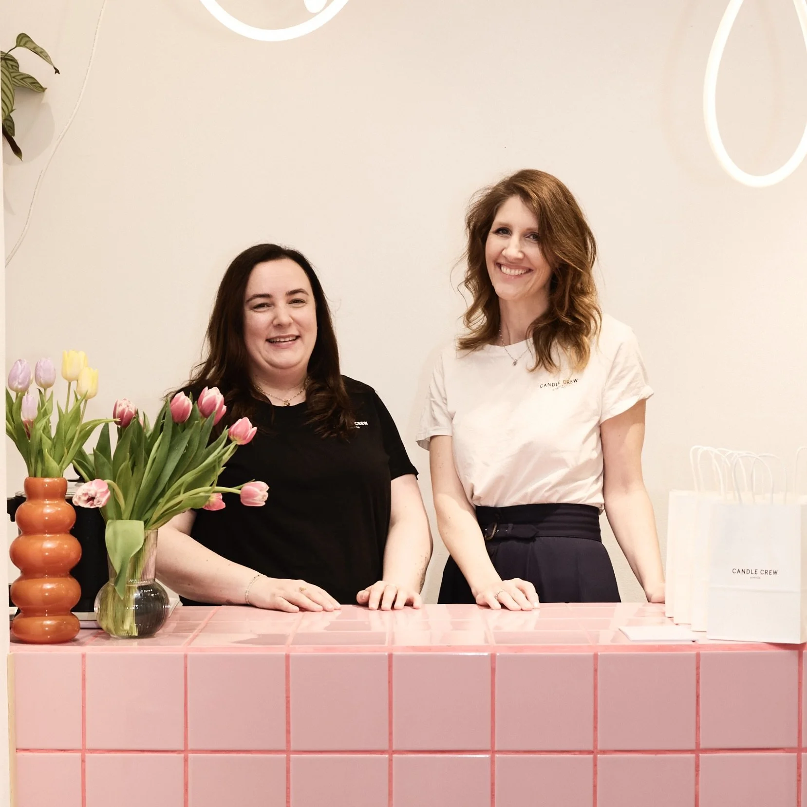 Zwei Frauen, die an einem pinkfarbenen Tisch stehen, halten sich an den Händen und lächeln. Vor ihnen sind Vasen mit Tulpen in verschiedenen Farben. Im Hintergrund ist eine weiße Wand mit dezenten Lichtern und einem Pflanzen.