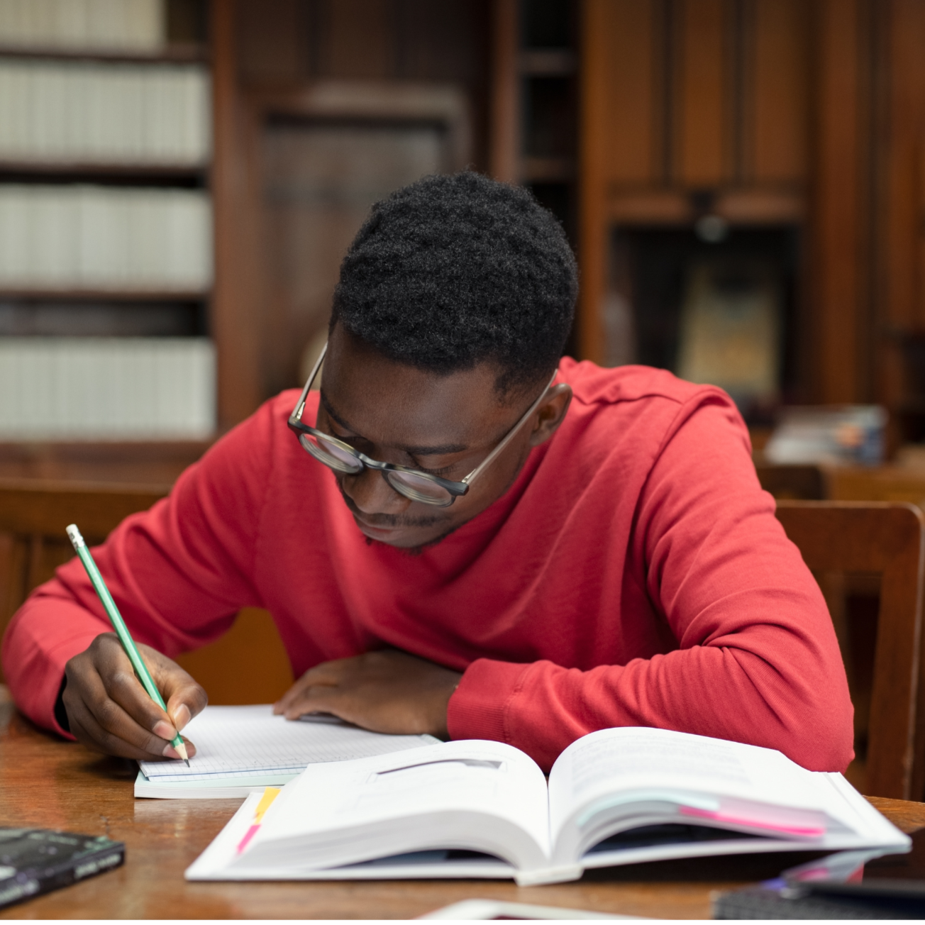 A young man with glasses in a red shirt writing in a notebook at a wooden table, with an open book and a bookshelf in the background.