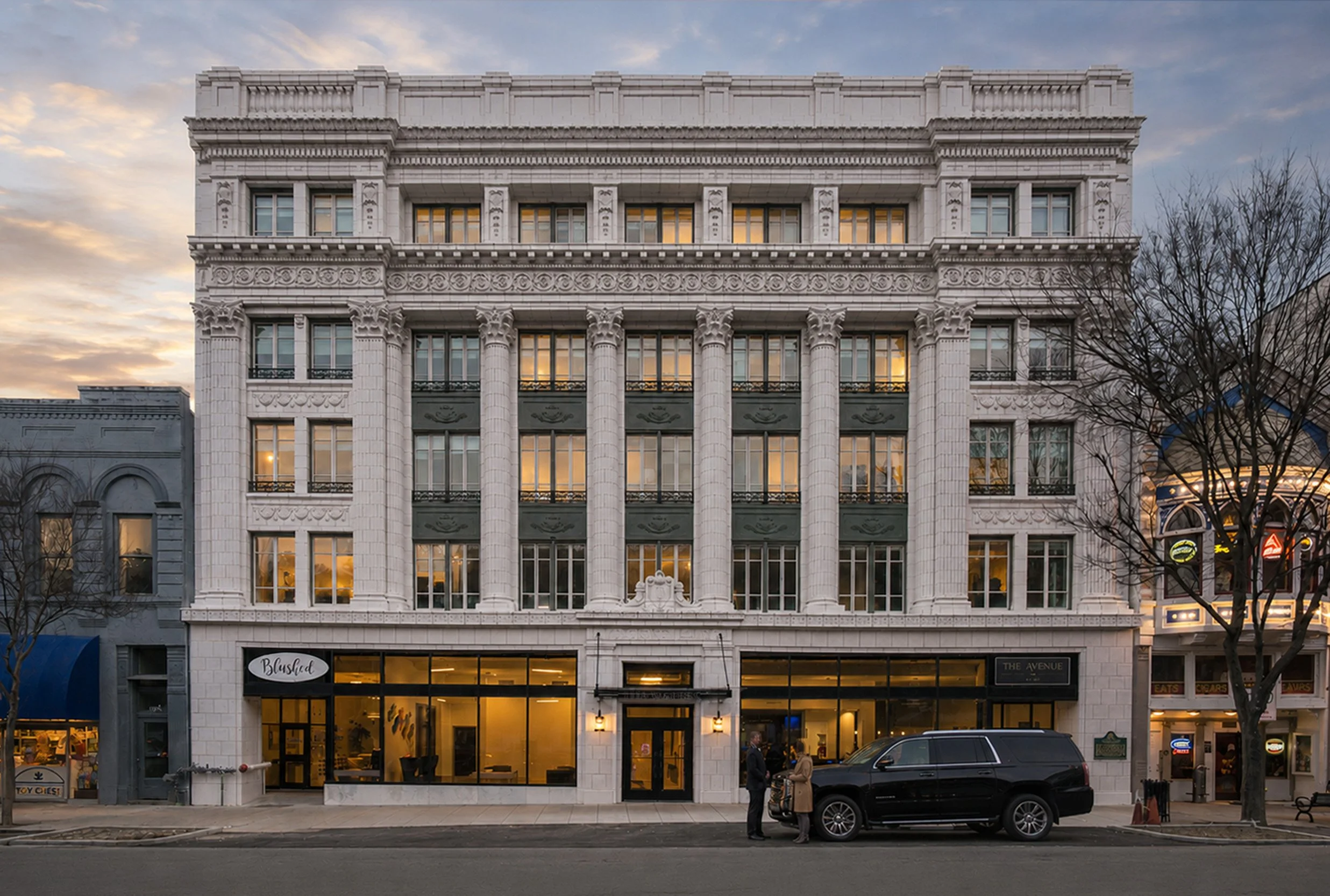 A multi-story building with ornate white architectural details, including columns and decorative moldings, during sunset. The storefront on the ground floor has large glass windows and is occupied by retail shops. There are leafless trees in front, and a few people and a parked black SUV are visible on the street.