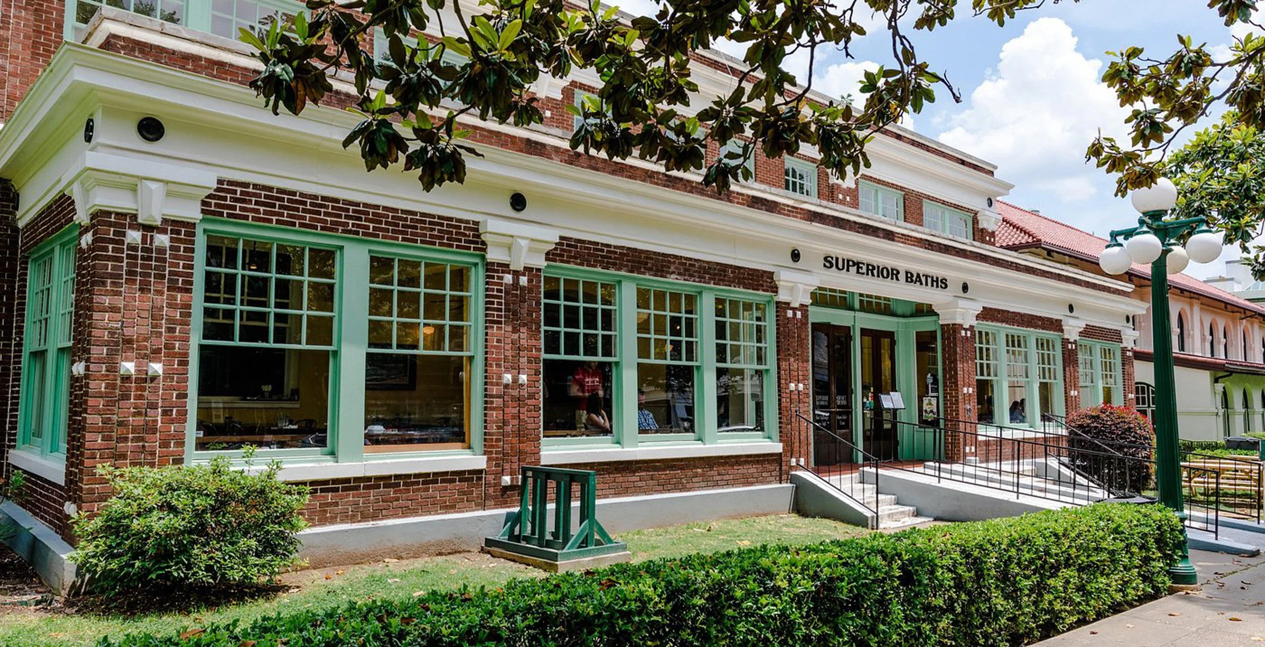 Exterior of a brick building with large green-framed window panes and a sign that reads "Superior Baths". There is a small set of stairs leading to the entrance, surrounded by trimmed bushes and a green lawn. A classic street lamp and some trees are 