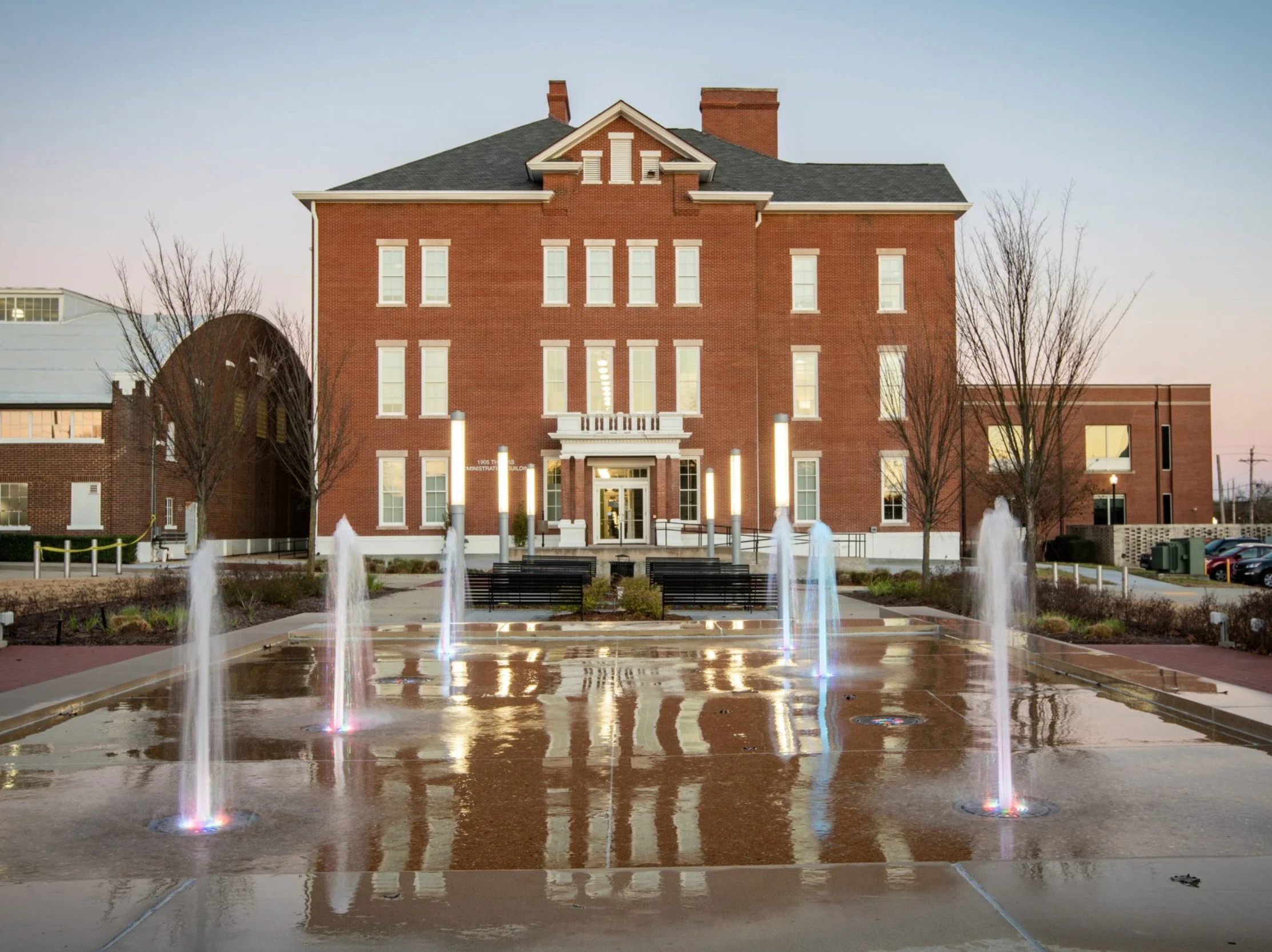 A large brick building with a peaked roof, multiple windows, and a small balcony entrance, situated behind a fountain with colorful lights, leafless trees, and parked cars, during dusk.