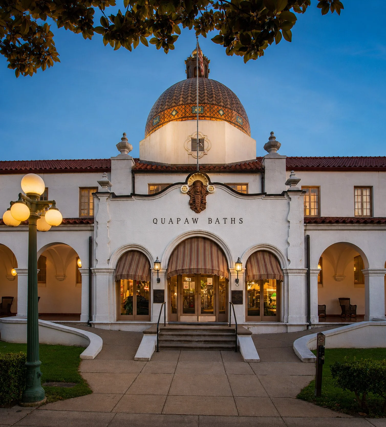 Front view of Quapaw Baths building with white walls, arched windows with striped awnings, and a tiled dome on top, set in evening lighting with trees and a street lamp nearby.