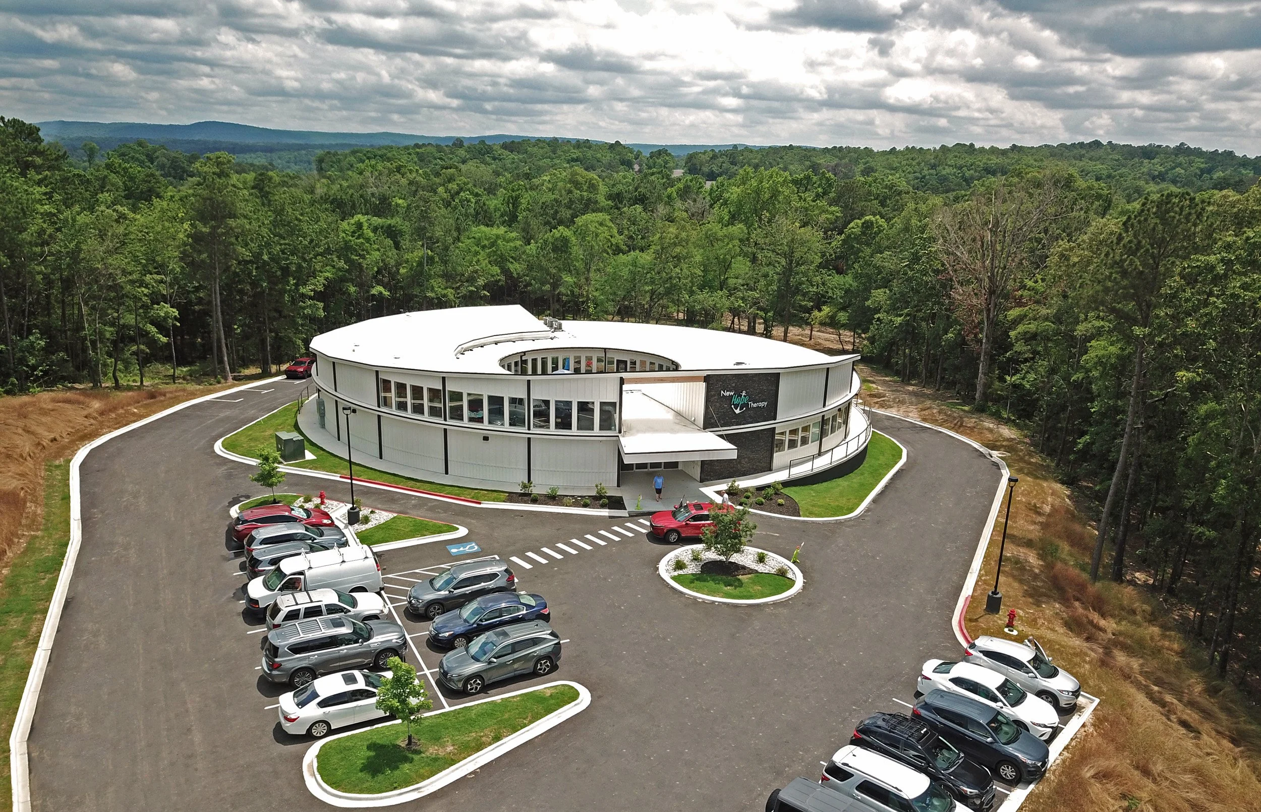 A modern, circular building with a white exterior and large windows, surrounded by a parking lot with cars, trees, and a cloudy sky in the background.