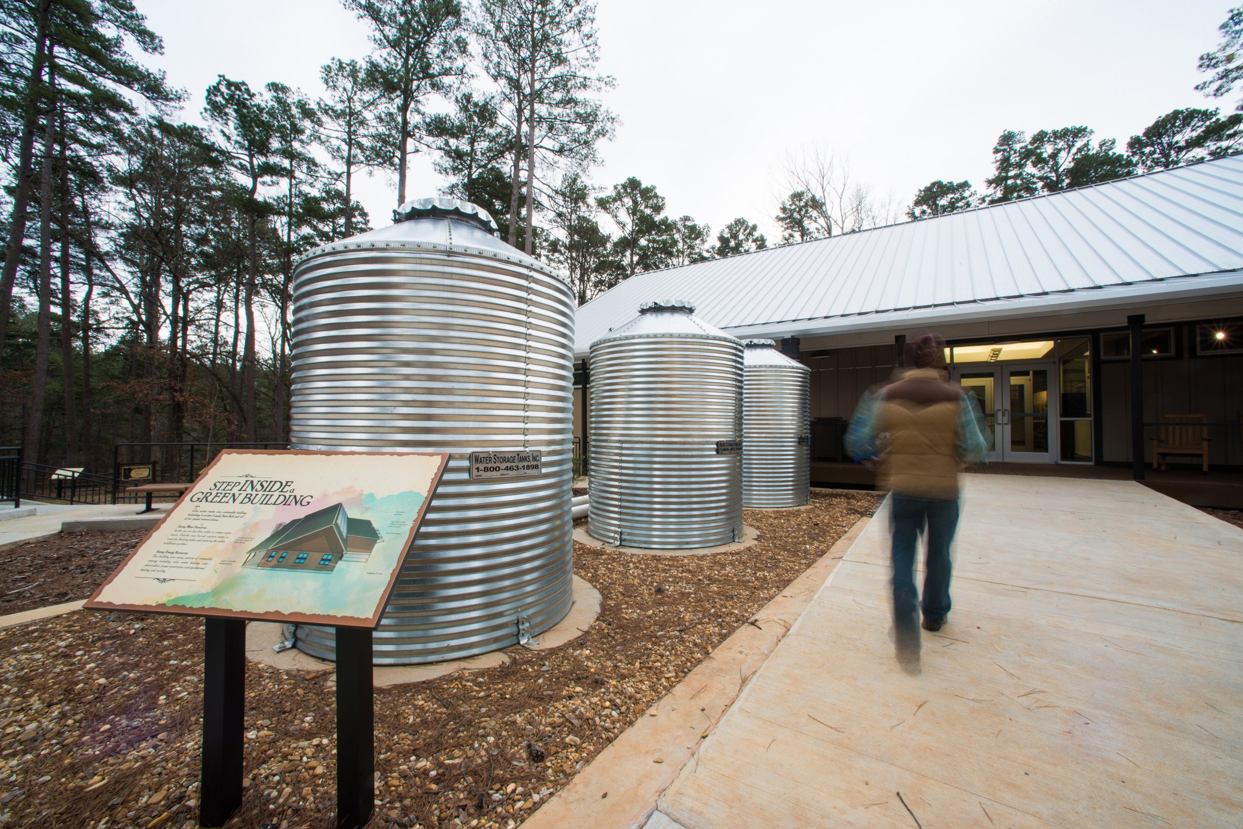 Metal water storage tanks outside a building surrounded by trees, with a person walking by and an informational sign about the building's green features.