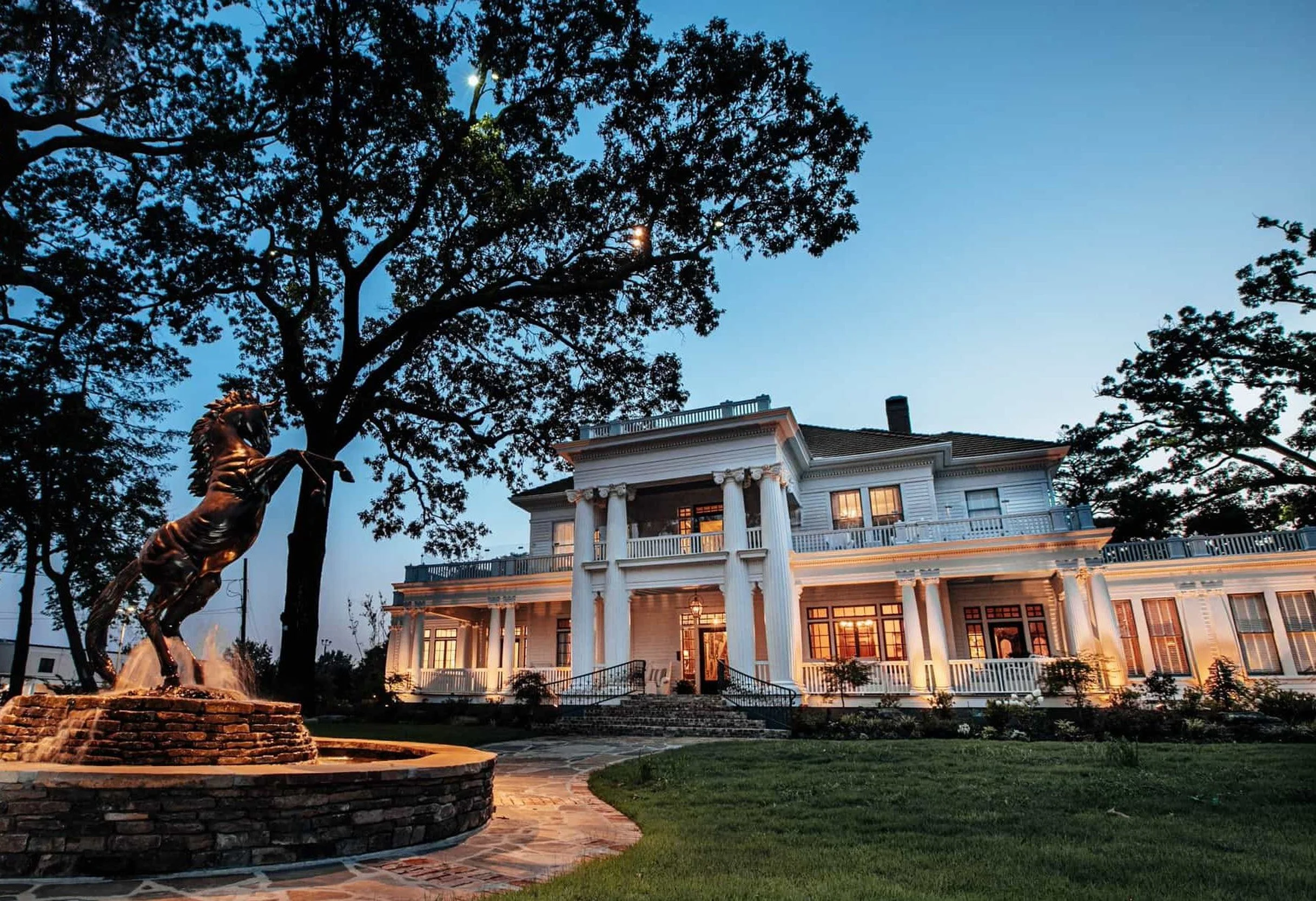A grand white house with a classic design featuring tall columns and a balcony, illuminated at dusk. In front, a statue of a horse and rider is visible near a fountain, with large trees framing the scene against a darkening sky.