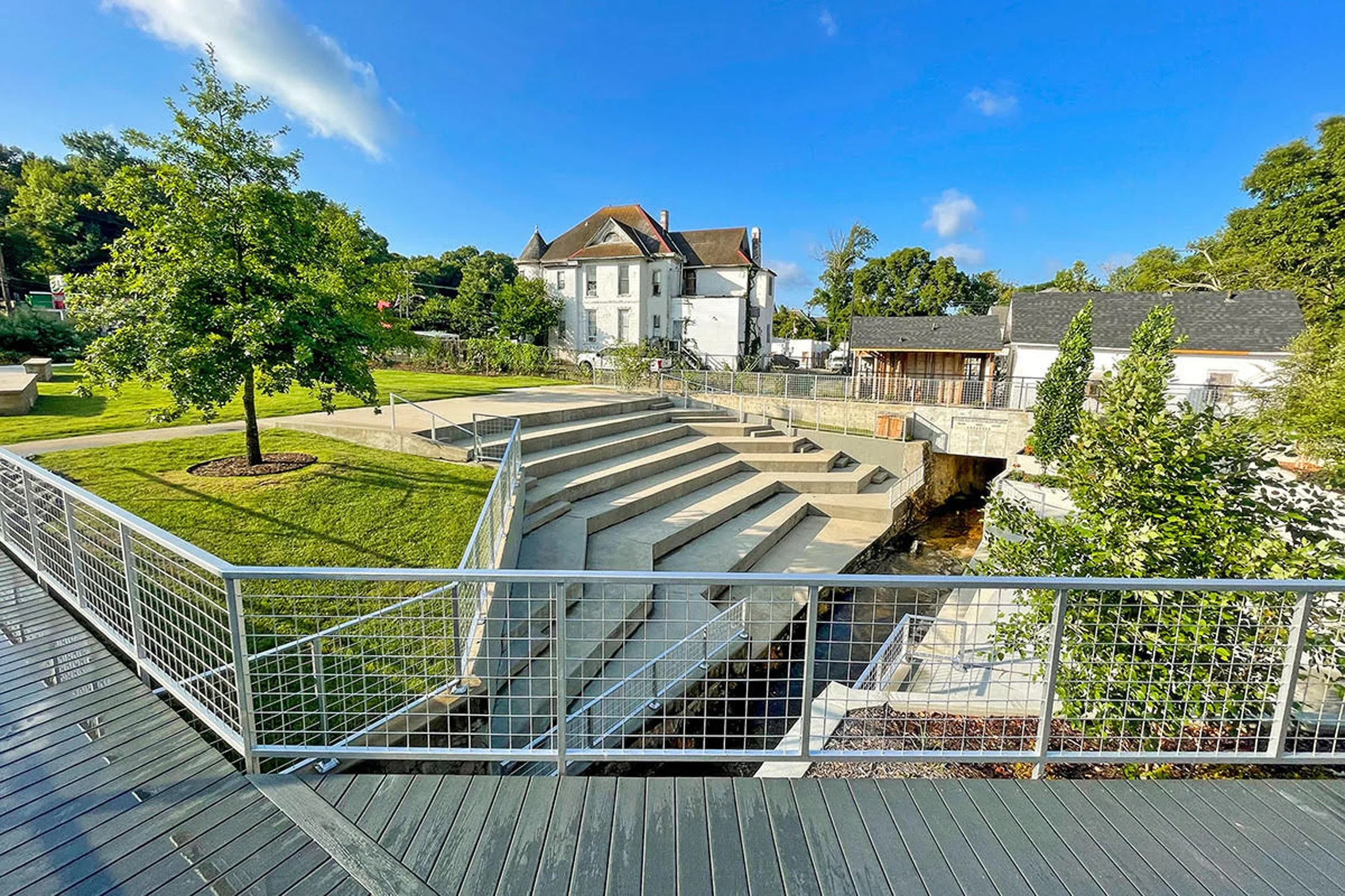 A landscaped backyard with a tree, concrete stairs, metal railing, and a grassy area. In the background are residential houses, trees, and a bright blue sky with a few clouds.