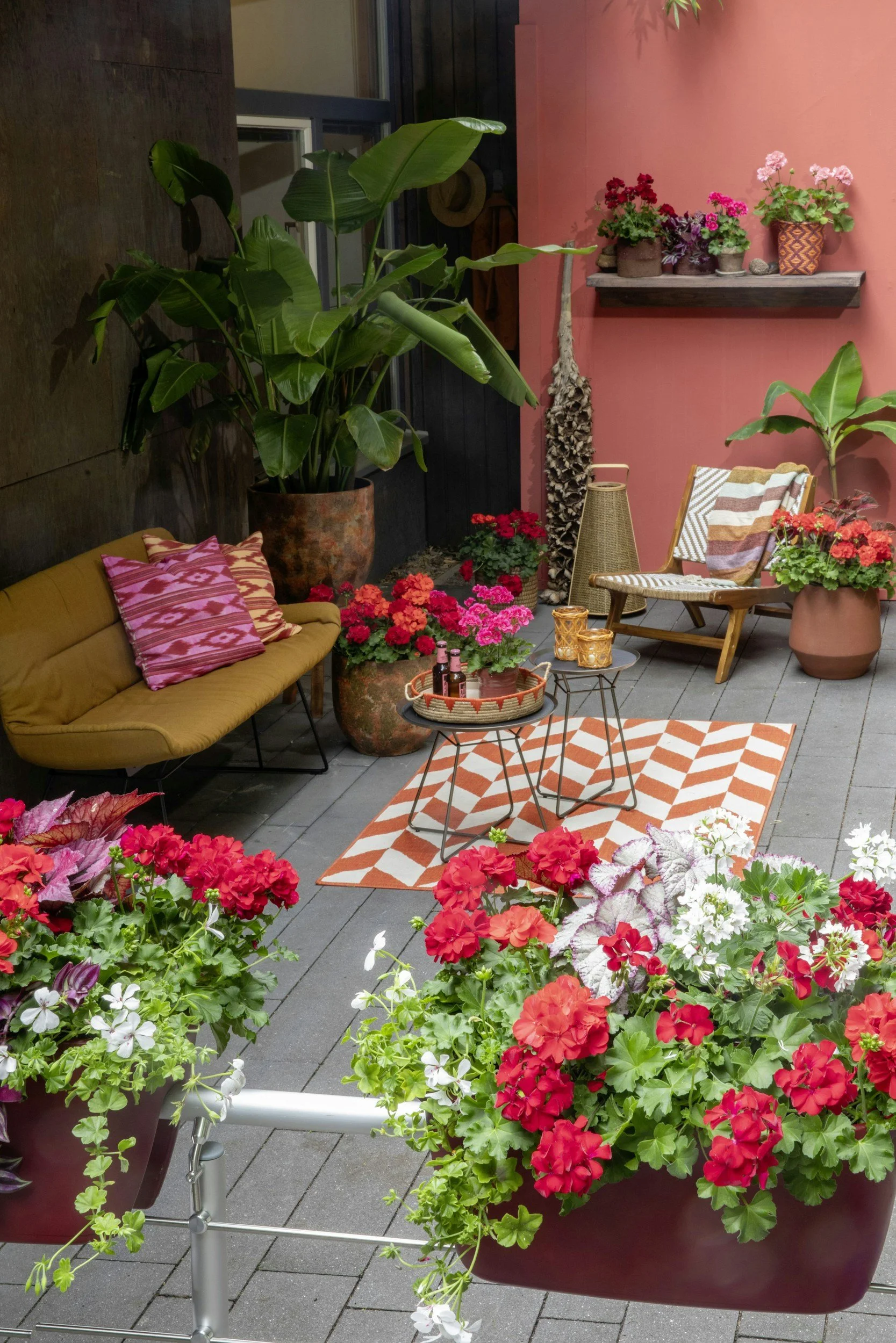 Bunte Blumen und Pflanzen auf einer Terrasse mit einem Gartenmöbel und Dekorationen in warmen Farbtönen.