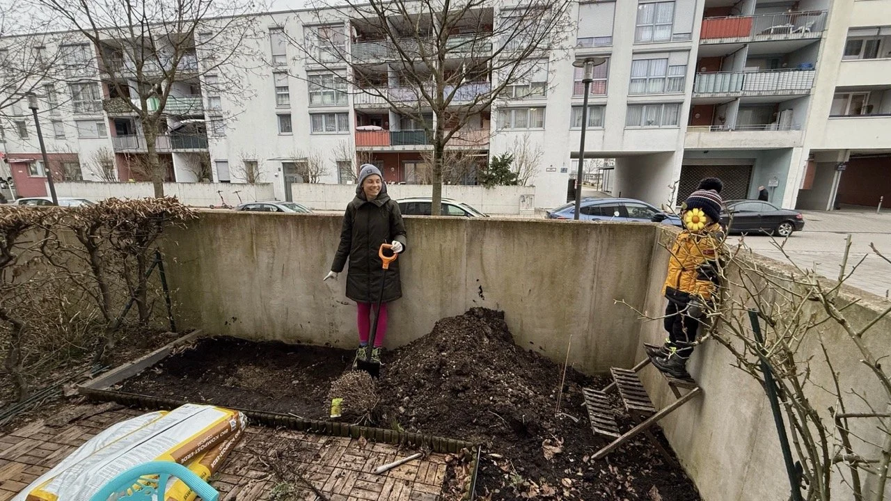 Zwei Personen pflanzen in einem kleinen Gartenbereich, mit Bauerngartengeräten und Materialien. Hintergrund mit Wohngebäuden.