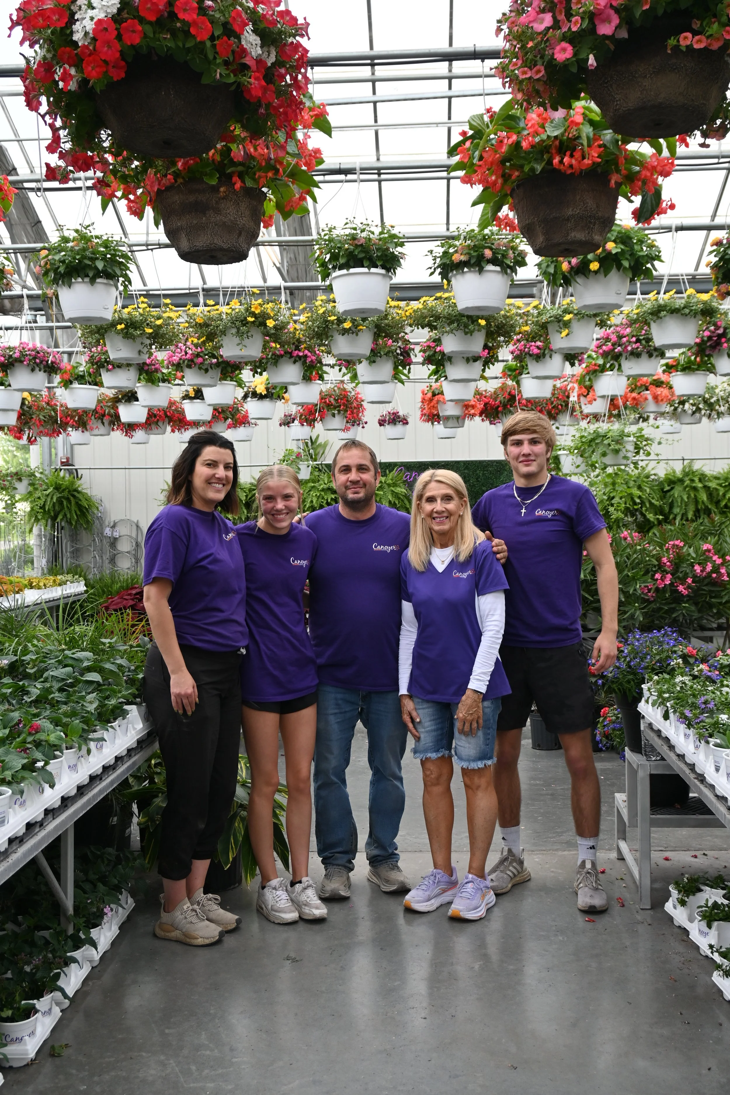 A group of five people pose together inside a greenhouse filled with hanging flower baskets and potted plants on shelves.