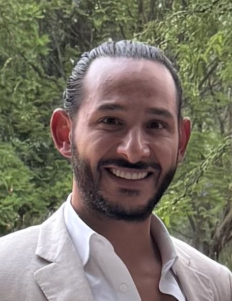 Smiling man with slicked-back hair and beard outdoors near trees