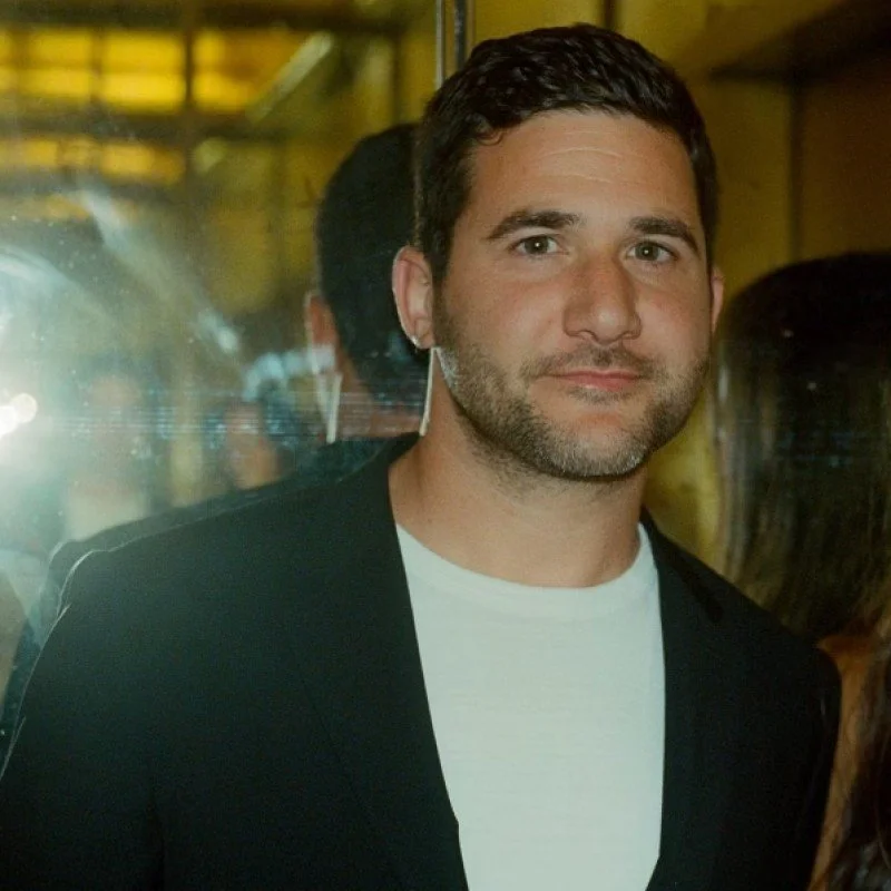 A man with short dark hair and a beard, wearing a white t-shirt and a black blazer, standing indoors with reflections and other people in the background.