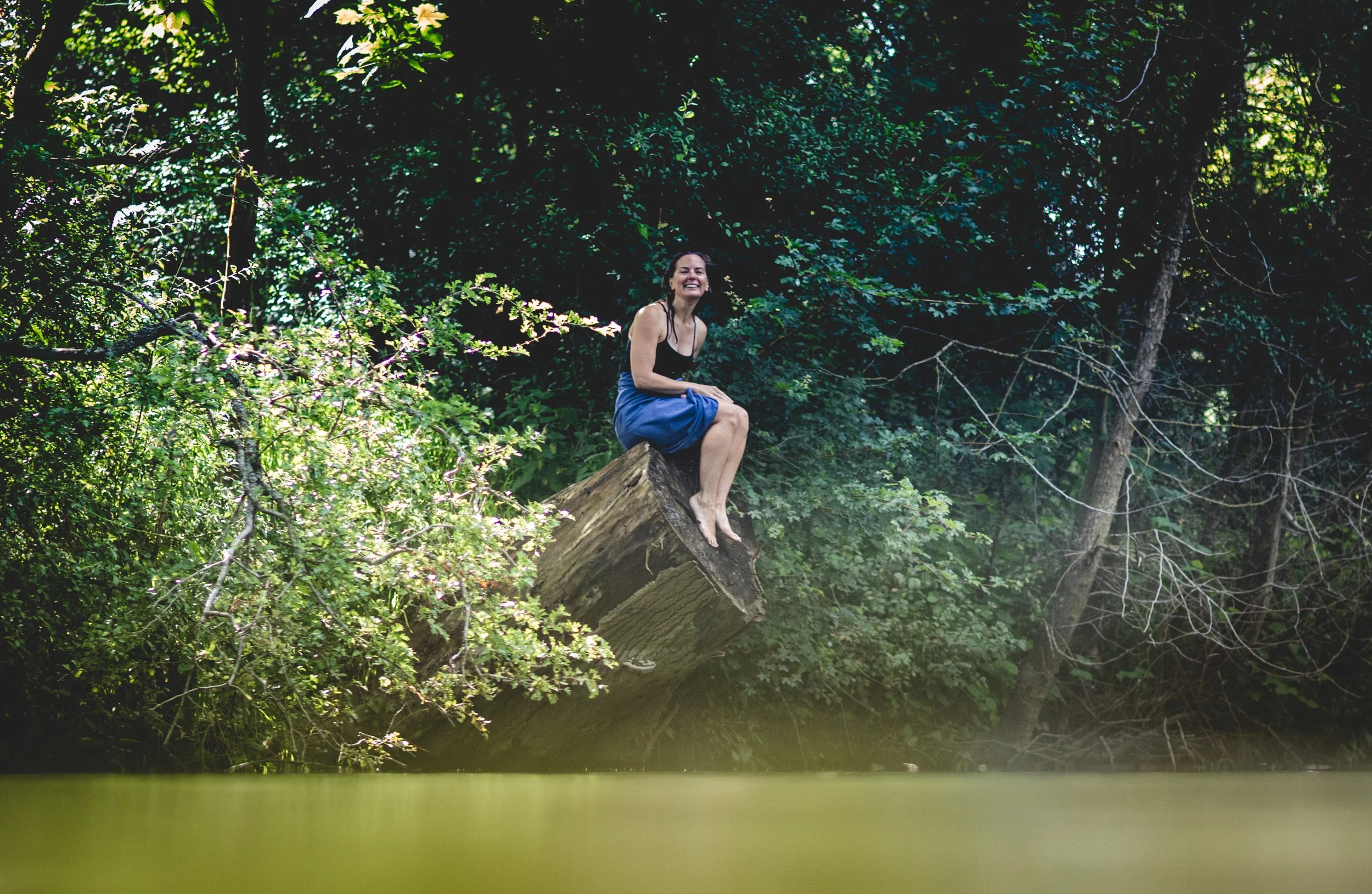 A woman sitting on a fallen tree trunk in a lush, green forest surrounded by dense foliage and trees.