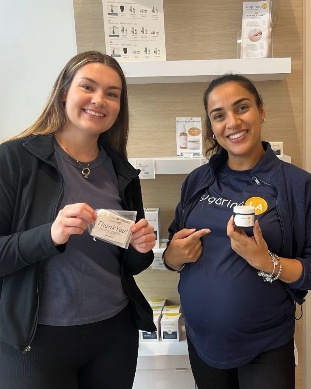 Two women smiling inside a store, one holding a thank you card and the other holding a small container of sugar. They are standing in front of shelves with skincare and health products.