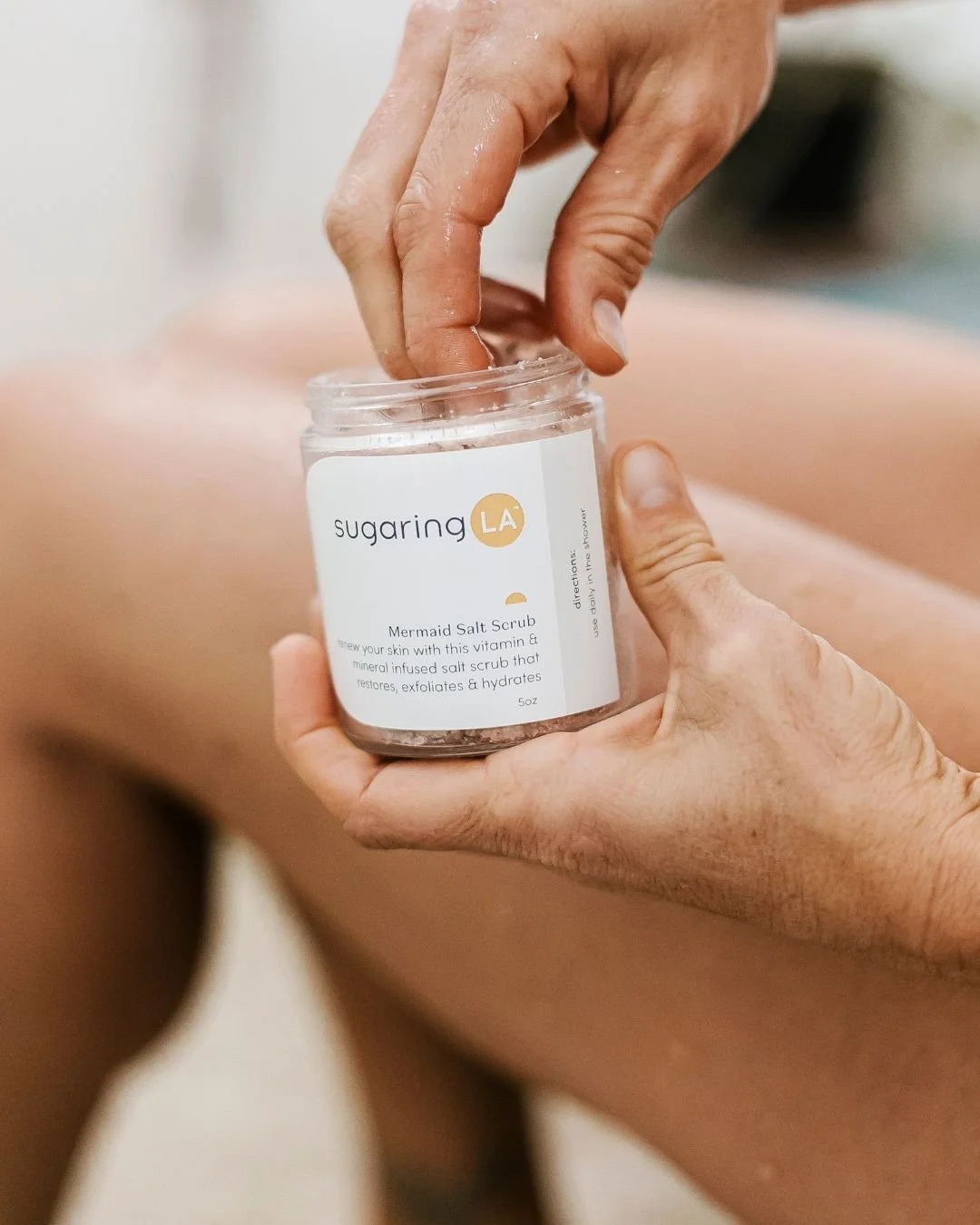 A person is holding a jar of Mermaid Salt Scrub from sugaring LA, with a pink-skinned person in the background.