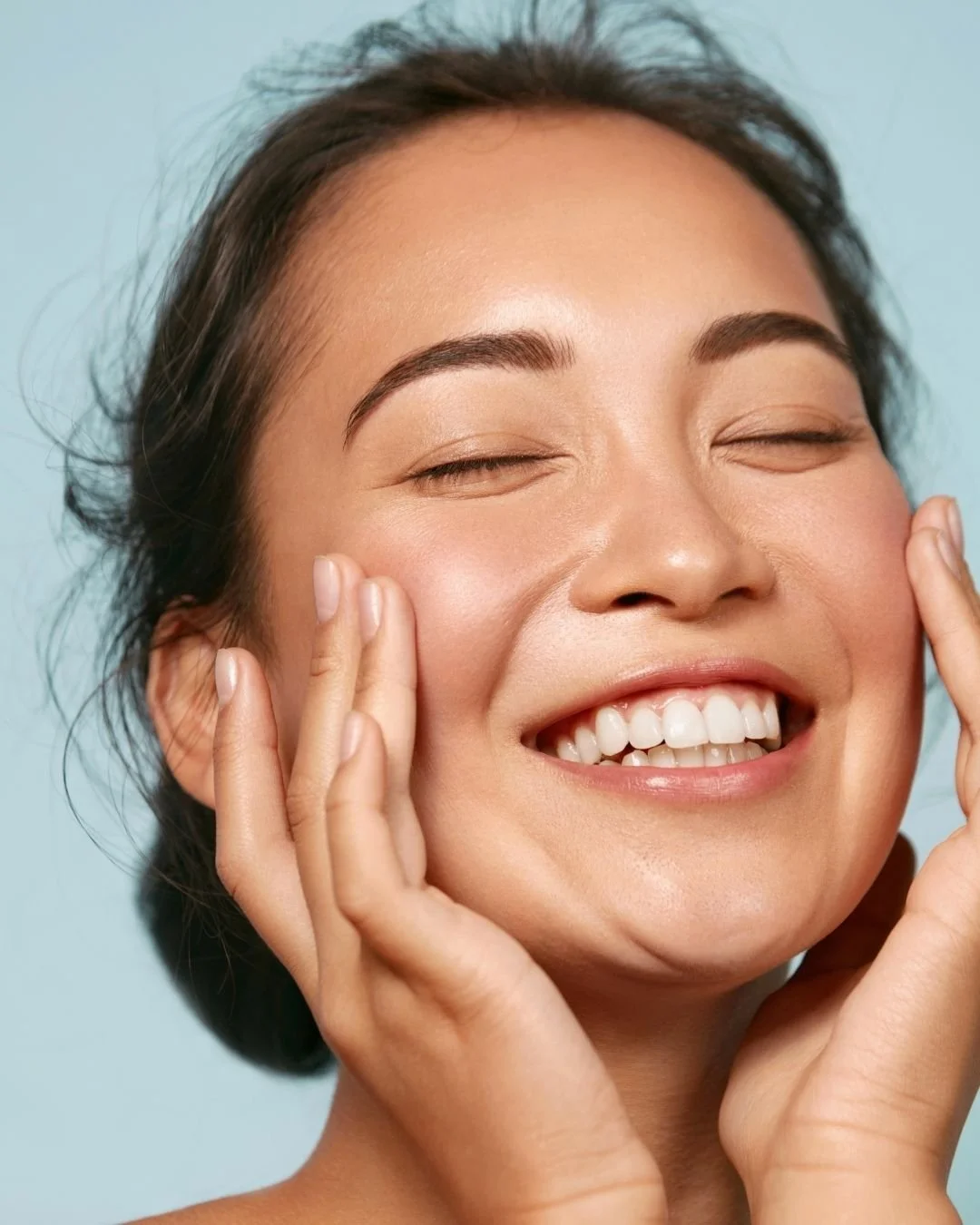 A woman smiling with eyes closed, touching her cheeks with both hands, showing clear skin and white teeth.