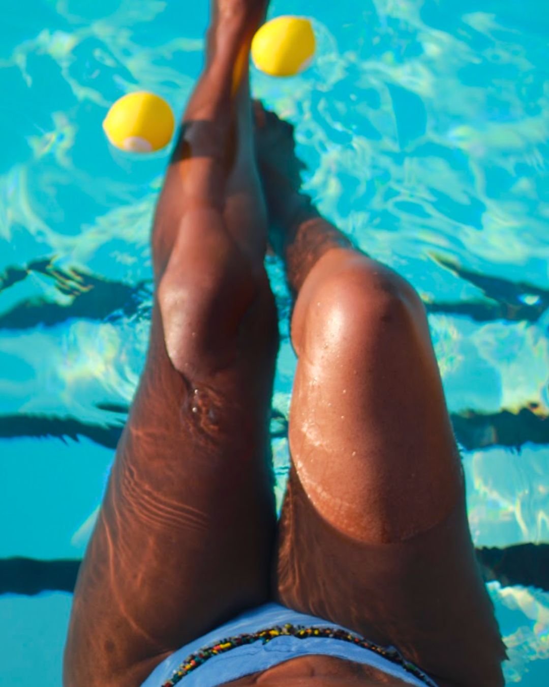 Person sitting on a pool float with their legs extended, two yellow pool balls resting on their legs, in a swimming pool with clear blue water.