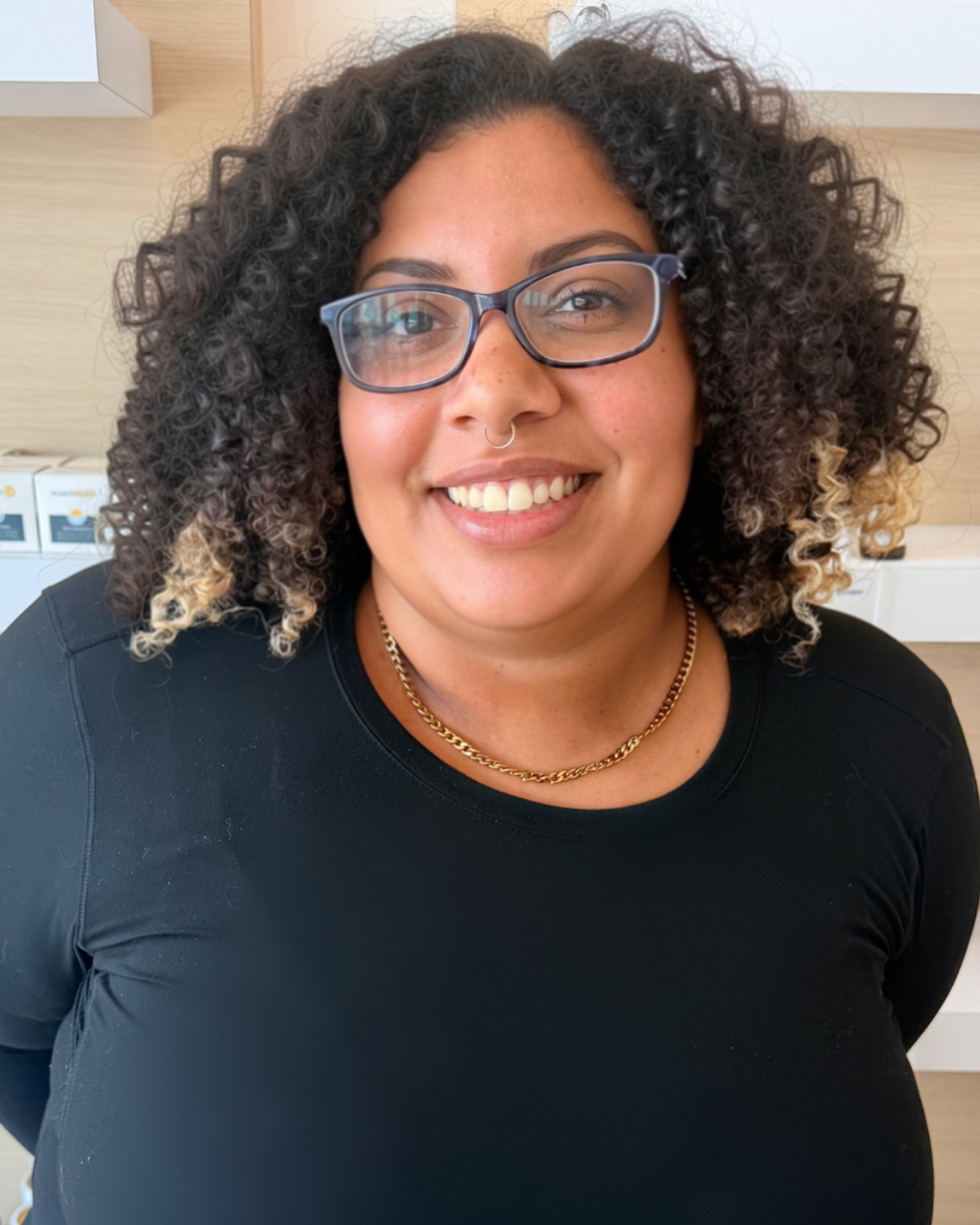 A woman with curly hair, glasses, and a septum piercing smiling indoors, wearing a black shirt and gold necklace.