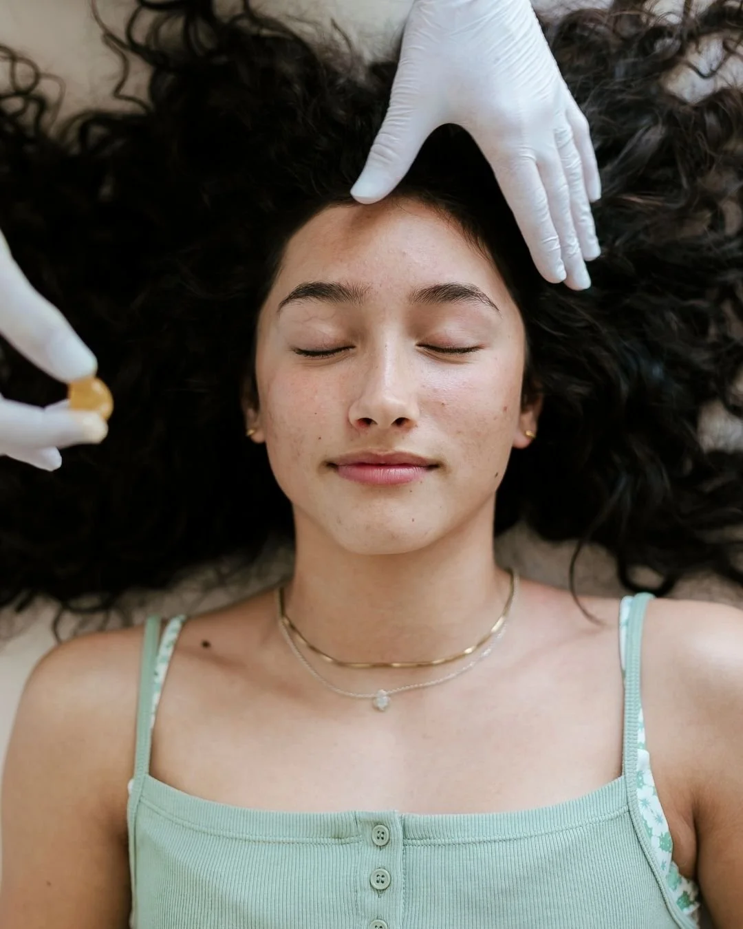 A woman with dark curly hair lying with her eyes closed while a person in white gloves applies a facial treatment or massage to her forehead.