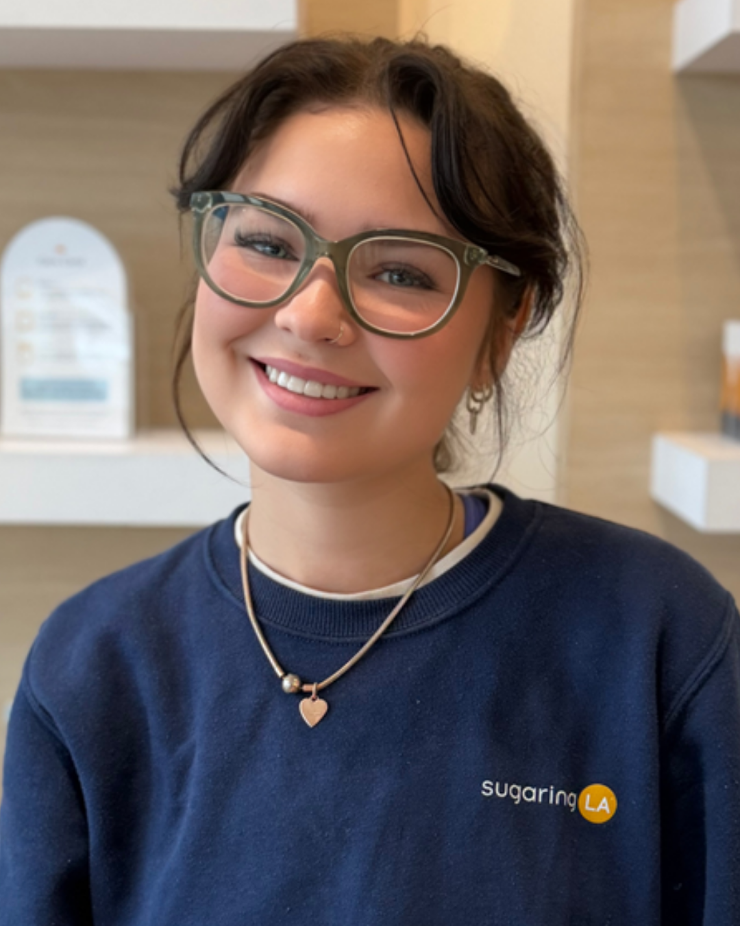 A smiling young woman with short dark hair, wearing large glasses, a navy blue sweatshirt with 'sugaring LA' logo, and a heart-shaped necklace, sitting indoors.