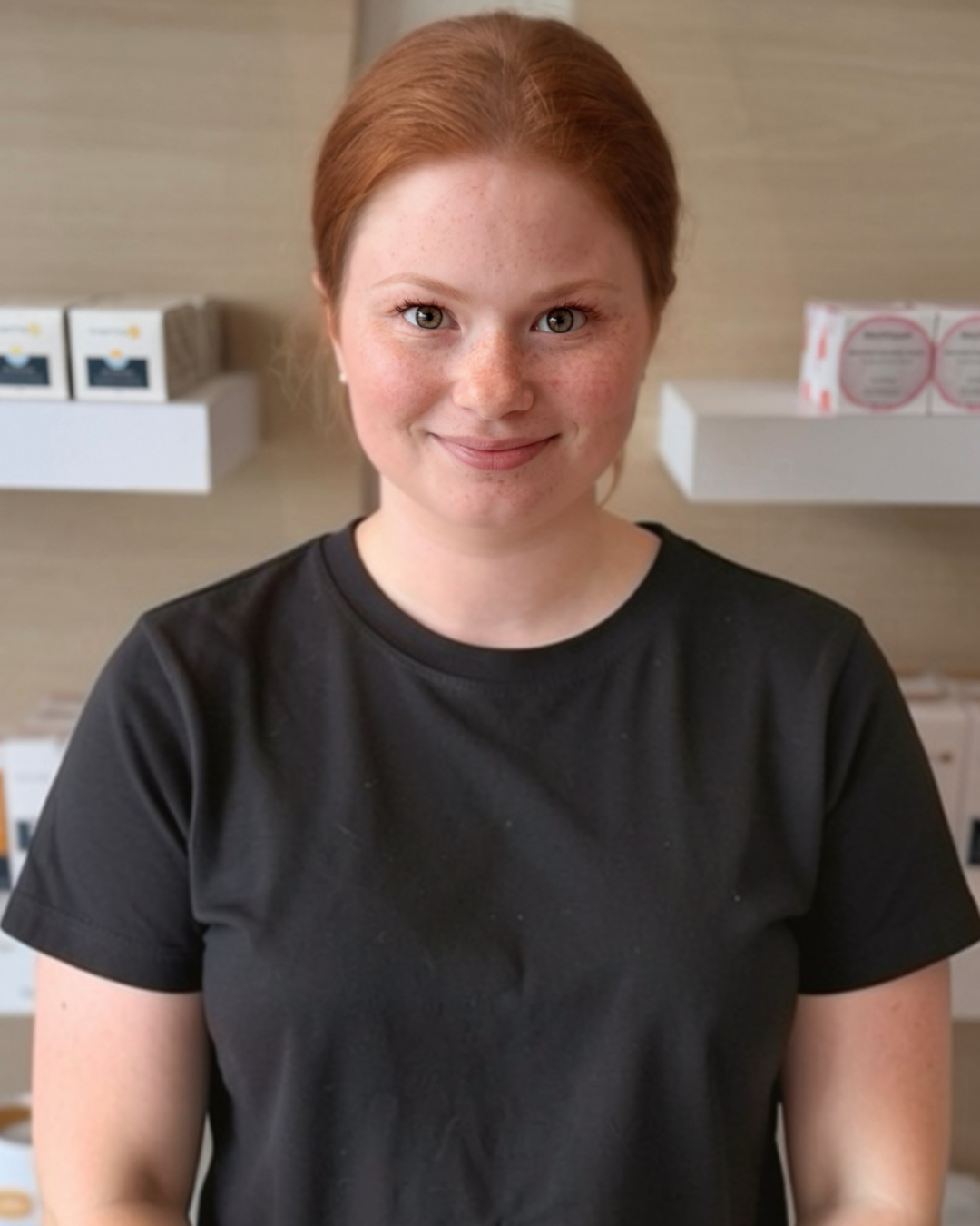 A young woman with red hair and freckles wearing a black t-shirt, smiling in front of retail shelves with boxes.