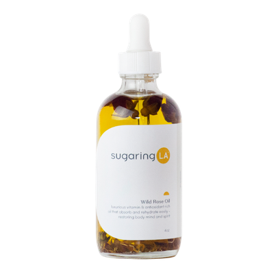A clear glass dropper bottle labeled 'sugaring LA Wild Rose Oil' containing yellow oil and dark rose petals inside.
