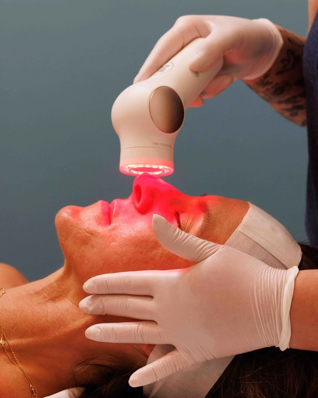 A person receiving a facial treatment with a device emitting red light, while wearing a protective headband and gloves.