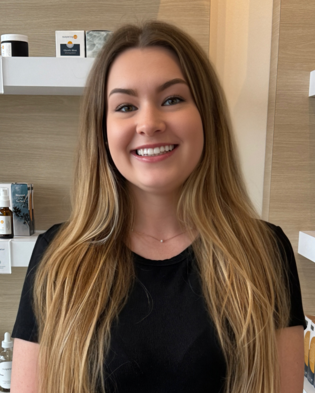 A woman with long, wavy light brown hair smiling indoors, wearing a black top and a delicate necklace, with shelves and skincare products in the background.