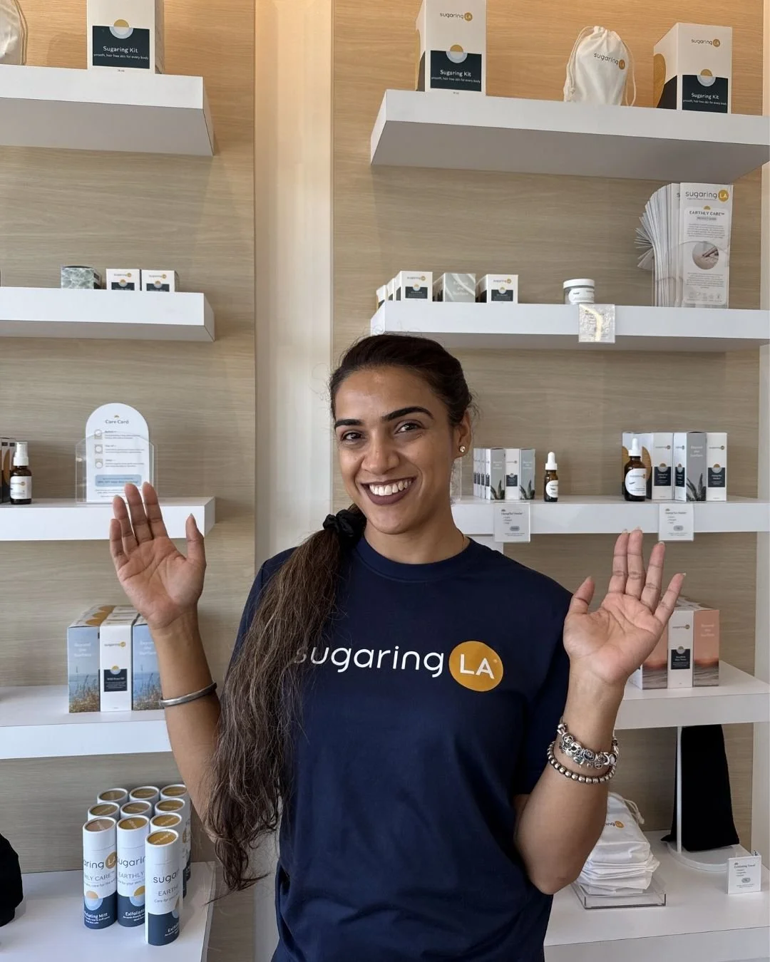 Woman smiling and raising both hands in front of shelves filled with health and wellness products, wearing a navy blue T-shirt with the logo 'Sugaring LA'.