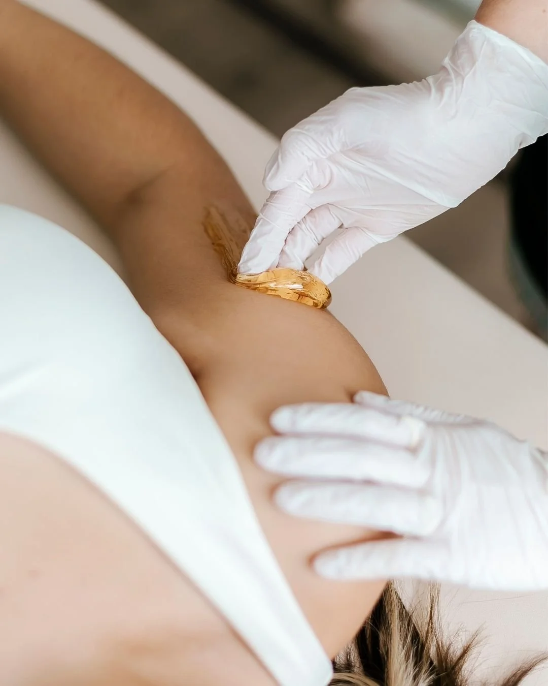 A person applying a golden-colored wax strip on their armpit, with a gloved hand pressing down, in preparation for hair removal.