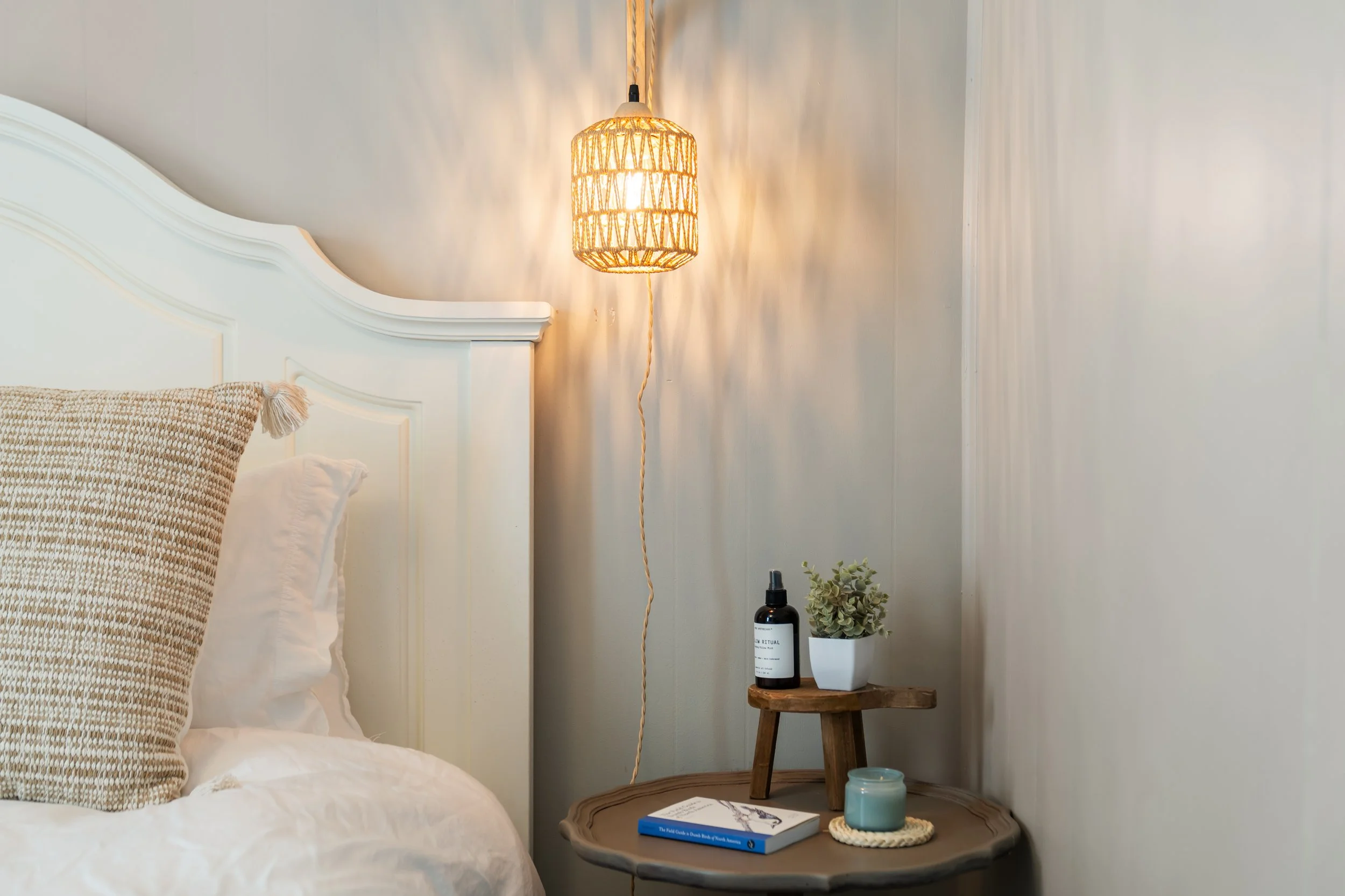Close-up of a cozy bedroom corner featuring a white headboard, textured beige pillow, hanging wicker lampshade, small wooden side table with a potted plant, a bottle of essential oil spray, a teal candle, and a book titled 'The Field Guide to Beach Body & Nook Animals'.