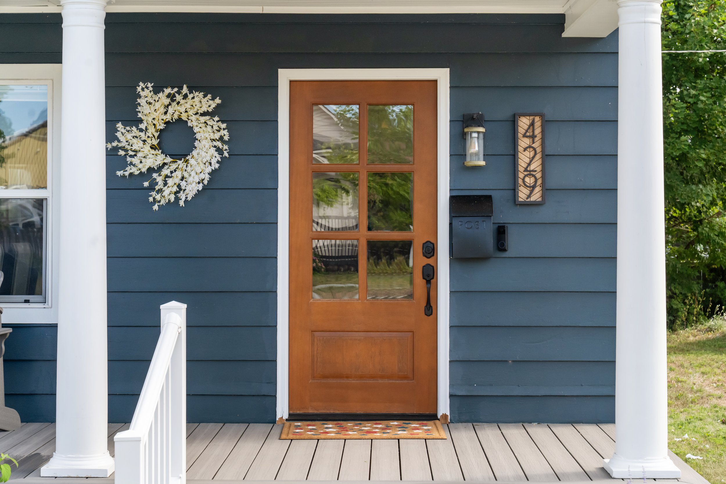 Front porch of a house with a wooden door, two large white columns, a decorative wreath on the left side, and wall-mounted outdoor light fixture on the right side. There is also a wooden house number plaque with the number 43 and a mailbox below it.
