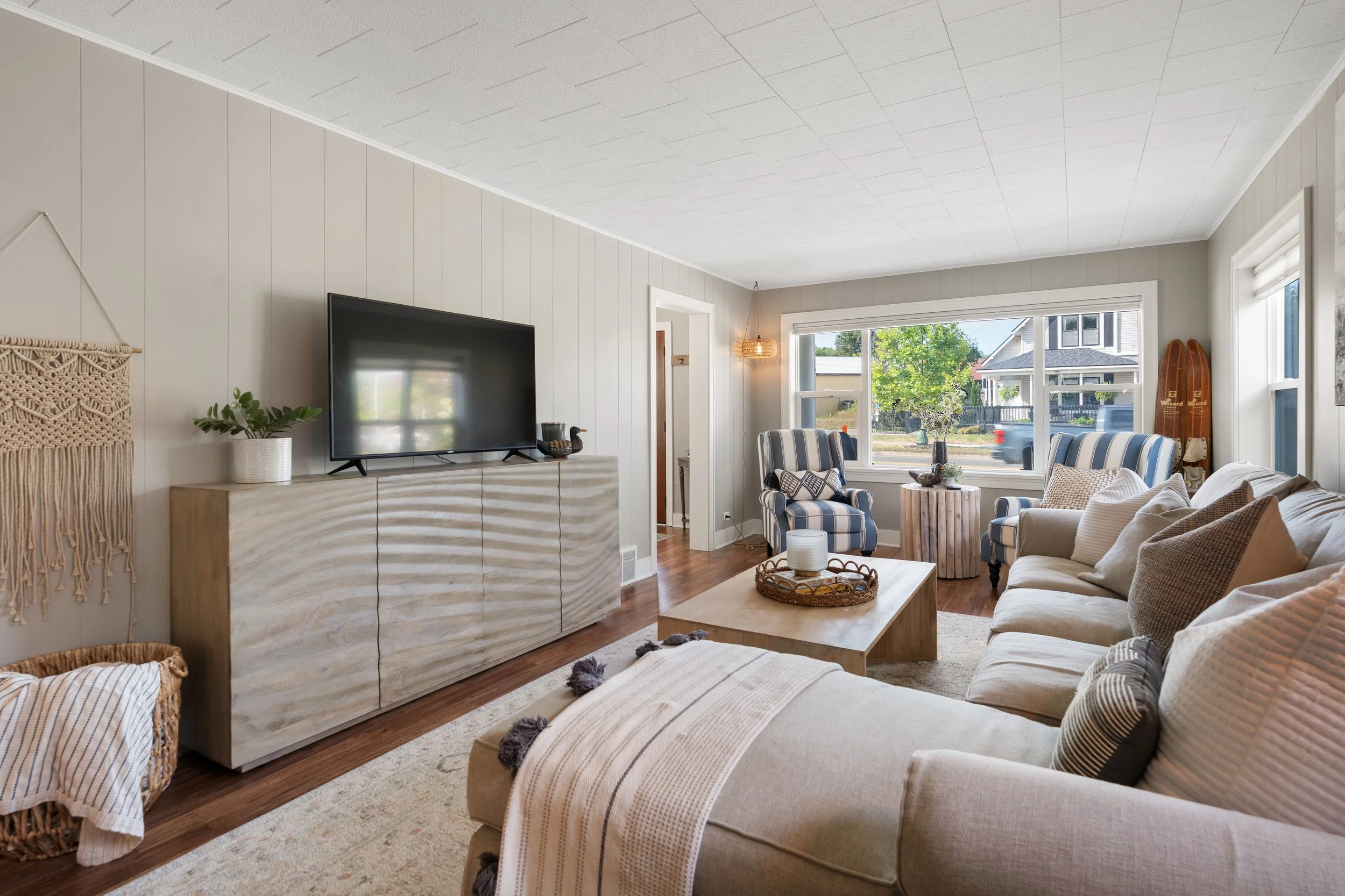 Living room with beige sofa, striped armchairs, wooden coffee table with tray, TV on a beige cabinet, windows showing a street view, and decor including plants and wall hanging.