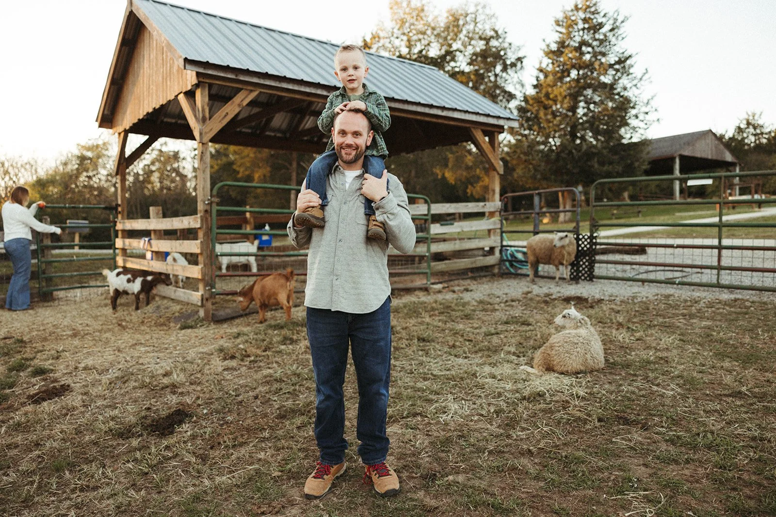 A man carrying a young boy on his shoulders at a farm with goats and sheep, with a fenced animal pen and trees in the background.