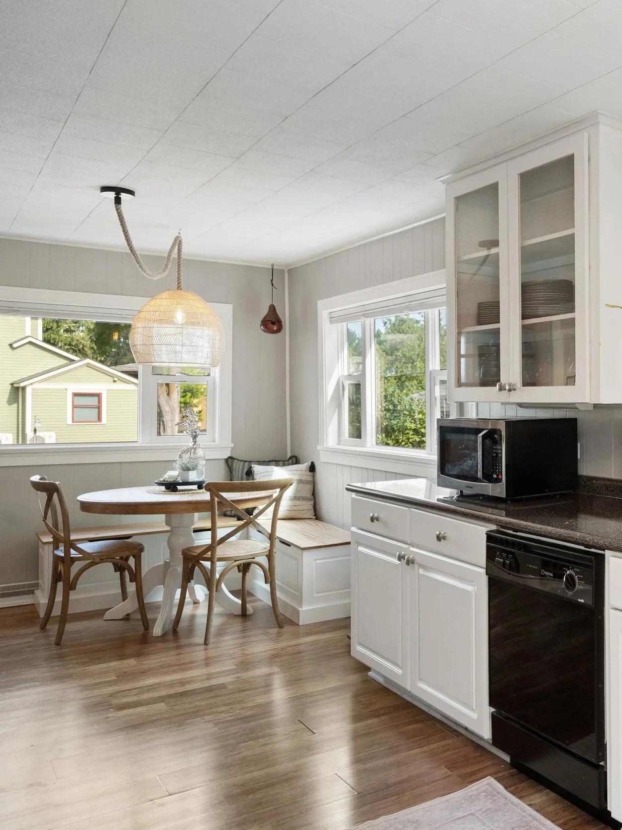 Bright kitchen with white cabinets, a microwave, and a black oven, next to a small wooden dining table with three chairs, and a large window showing a green backyard.