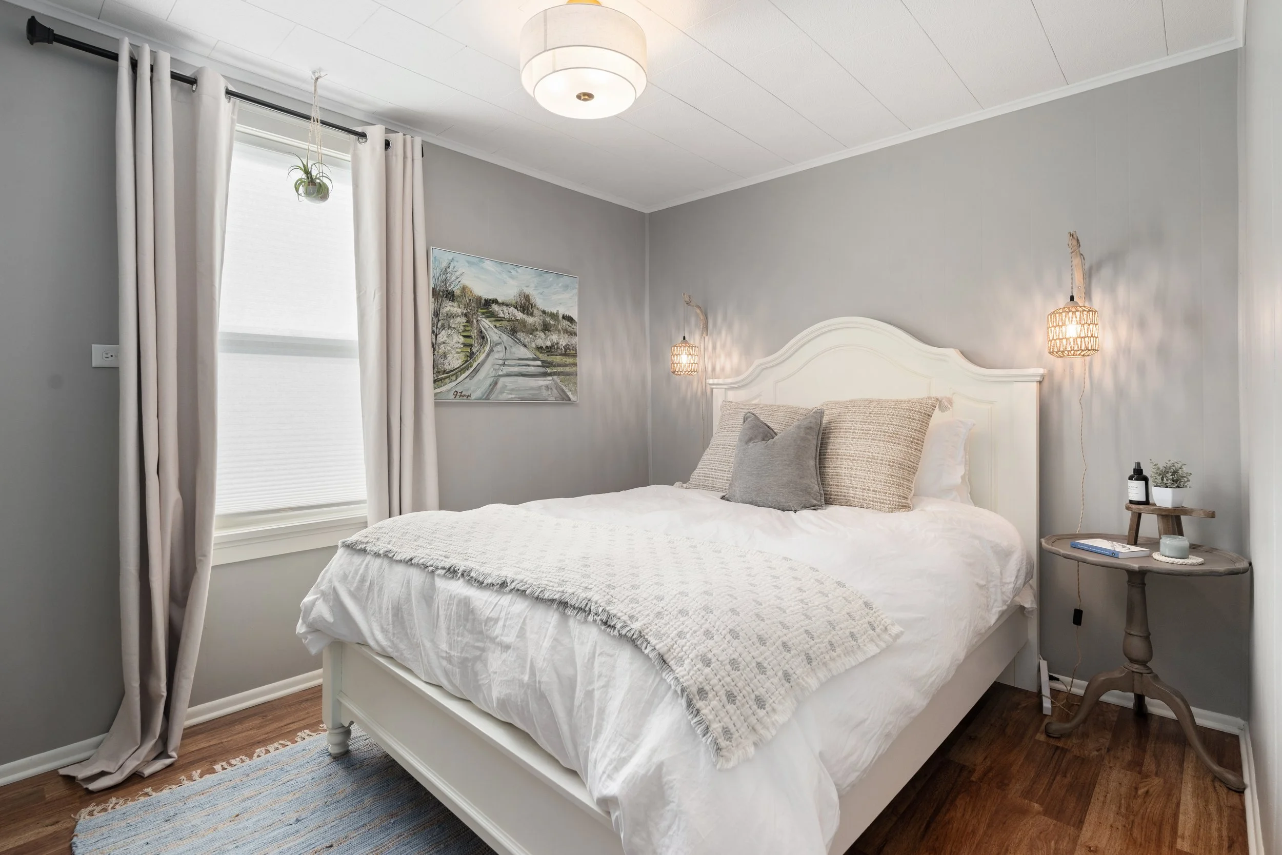 A cozy bedroom with a white bed, beige and gray pillows, gray walls, a painting of a countryside scene, a small bedside table with a book, candles, and a potted plant, natural light coming through a window with white curtains, and a ceiling light