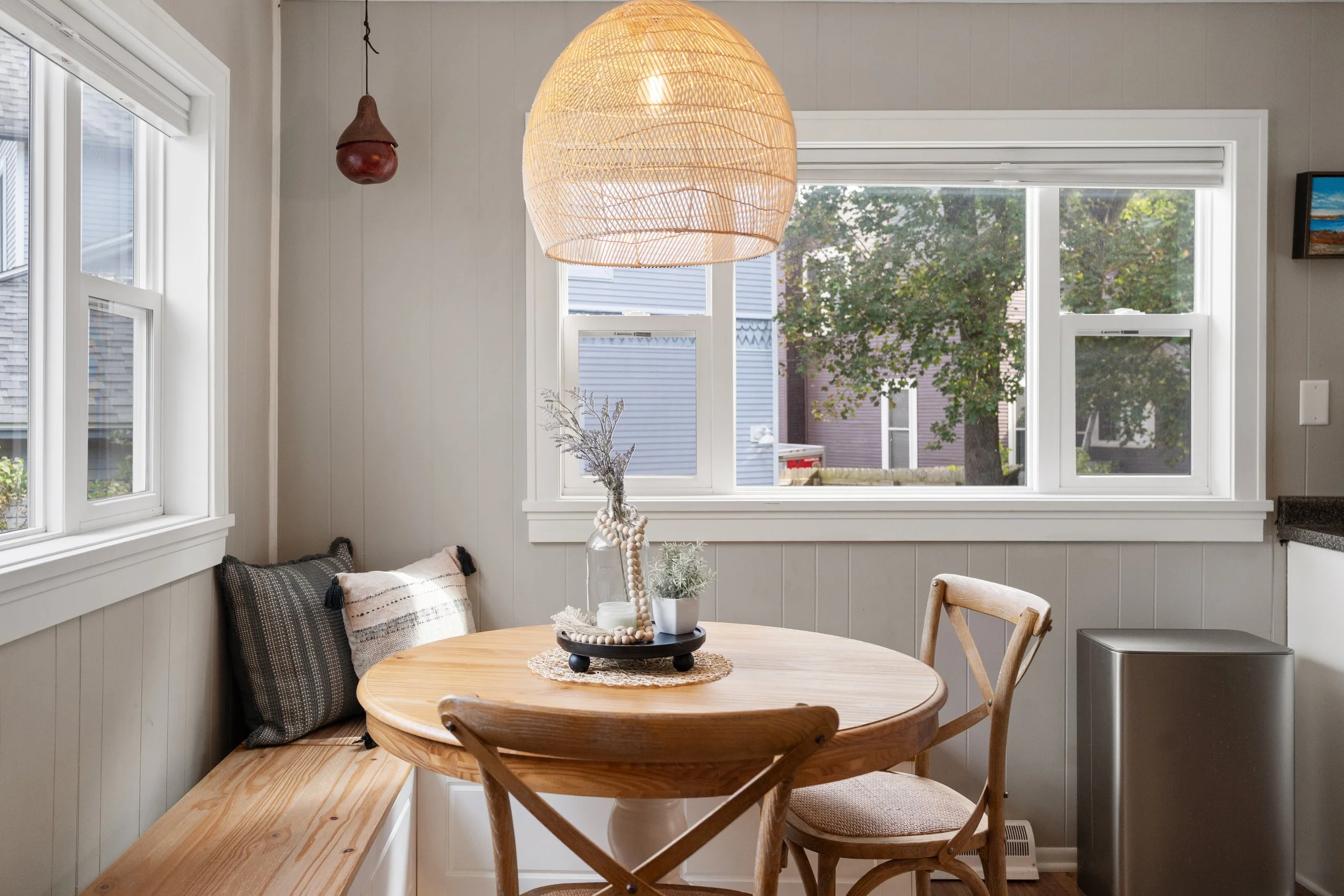 The image shows a cozy kitchen dining area with a round wooden table, two wooden chairs, and a built-in wooden bench with cushions. There is a decorative centerpiece on the table with a glass jar, beads, and plants. Large windows with white framing let in natural light and show trees outside. A woven pendant light hangs above the table, and a small wooden decorative object is on the window sill.
