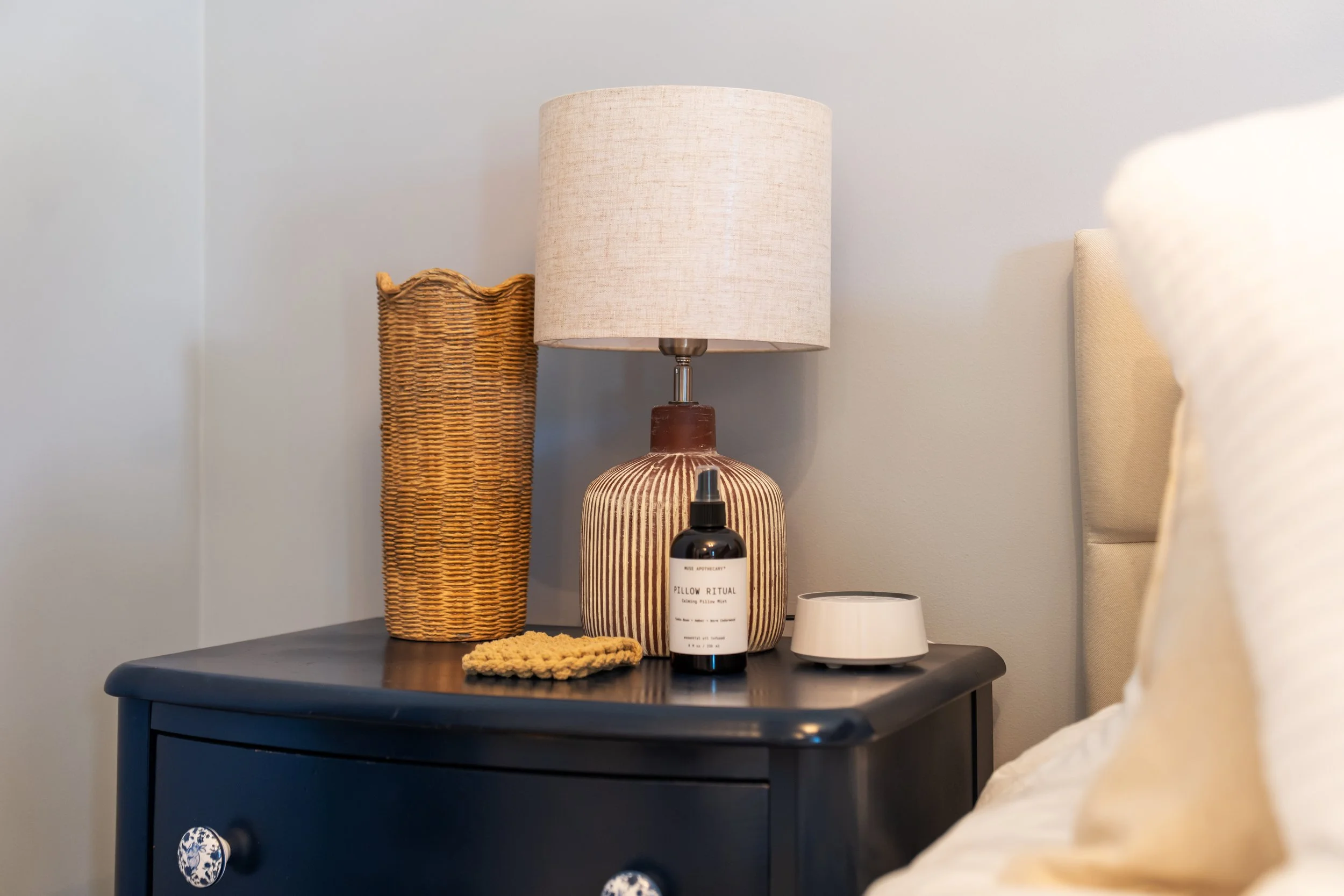 A dark blue bedside table with a lace knob. On top is a wicker basket, a table lamp with a beige shade, a striped ceramic vase labeled 'Pillow Ritual,' a small black dropper bottle, and a white container. A cream-colored bed with a cushion is visible to the right.