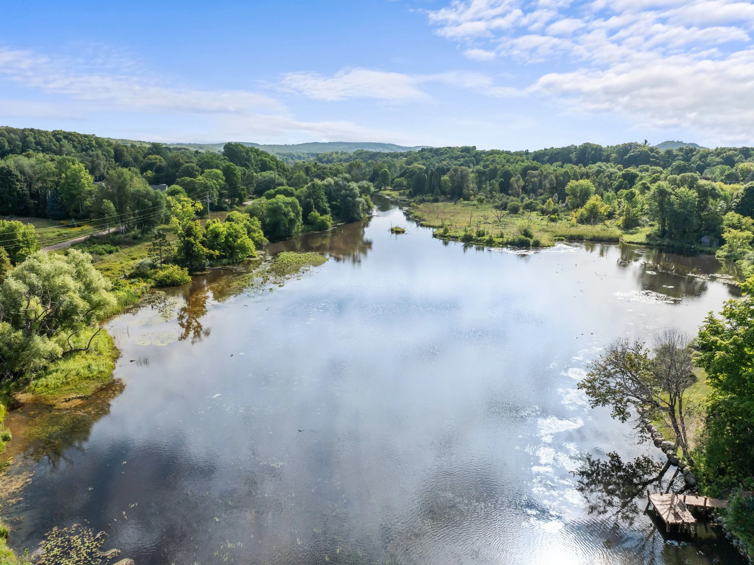 A scenic view of a river flowing through lush green trees and rolling hills under a partly cloudy sky.
