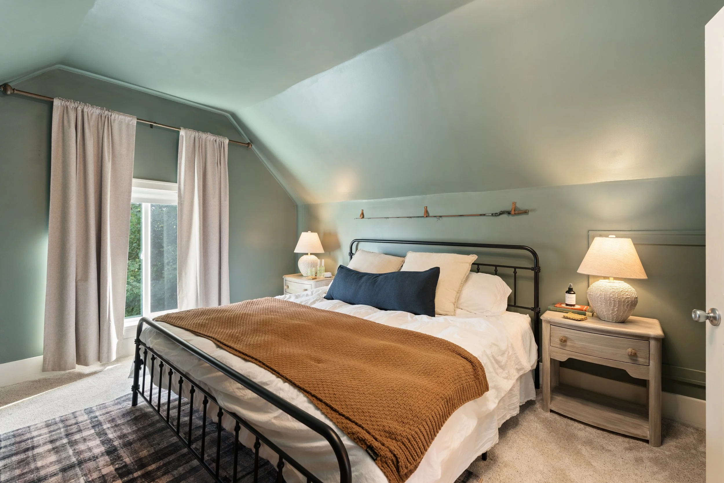A cozy bedroom with a black metal bed frame, beige and white bedding, and a brown knitted blanket. A wooden nightstand with a lamp, a bottle of essential oil, and books. Light green painted walls, a window with beige curtains, and a plaid area rug.