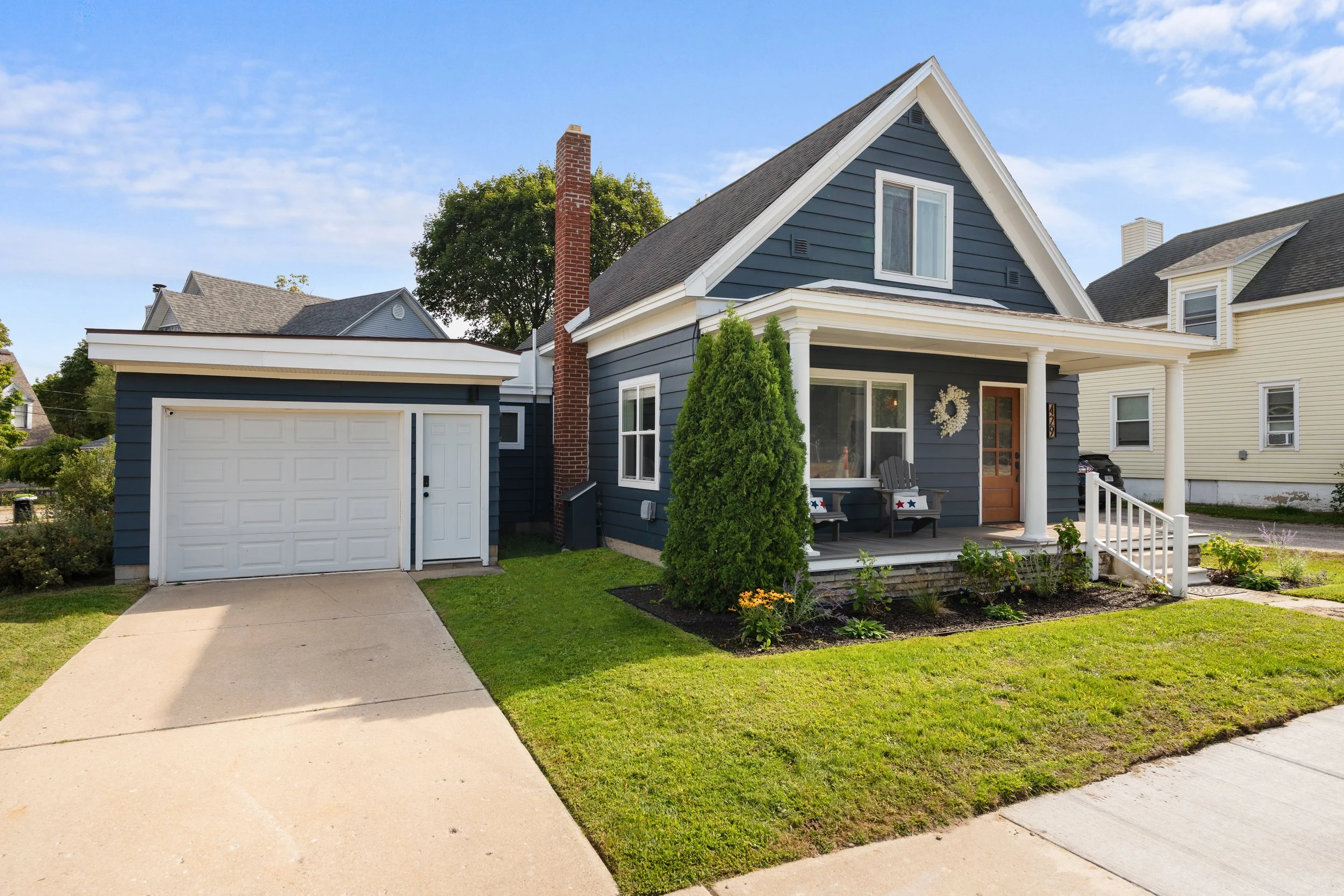 Blue house with white trim, porch with a wreath on the door, small seats with star decorations, and a green bush in front. Garage on the left, neighboring houses in the background, well-maintained lawn, and a clear blue sky.