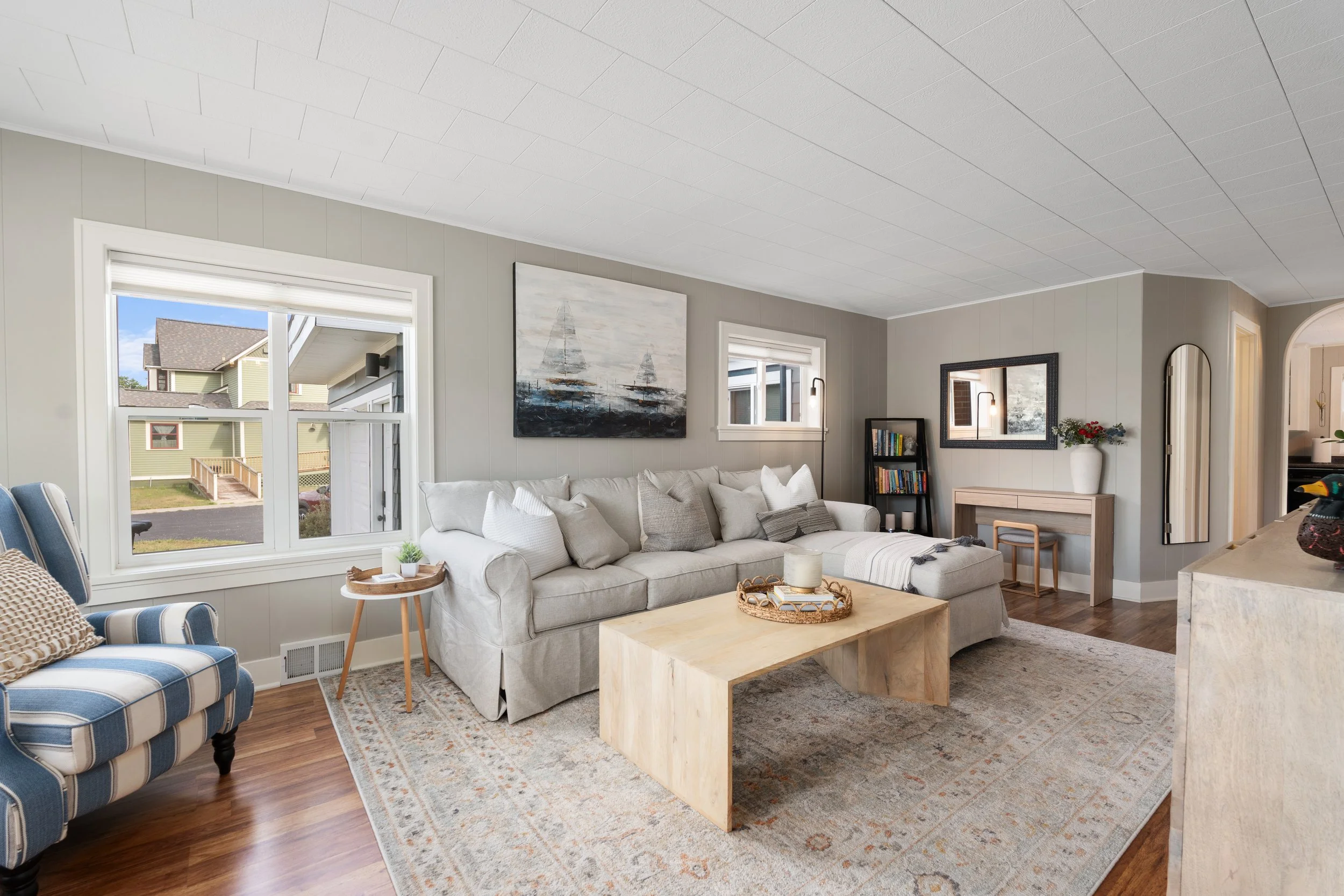 A cozy living room with a large beige sectional sofa, a wooden coffee table, and colorful chairs. The room has light gray walls, white trim, and large windows with a view of neighboring houses. Artwork and decorative items are displayed on the walls and shelves, creating a warm and inviting atmosphere.