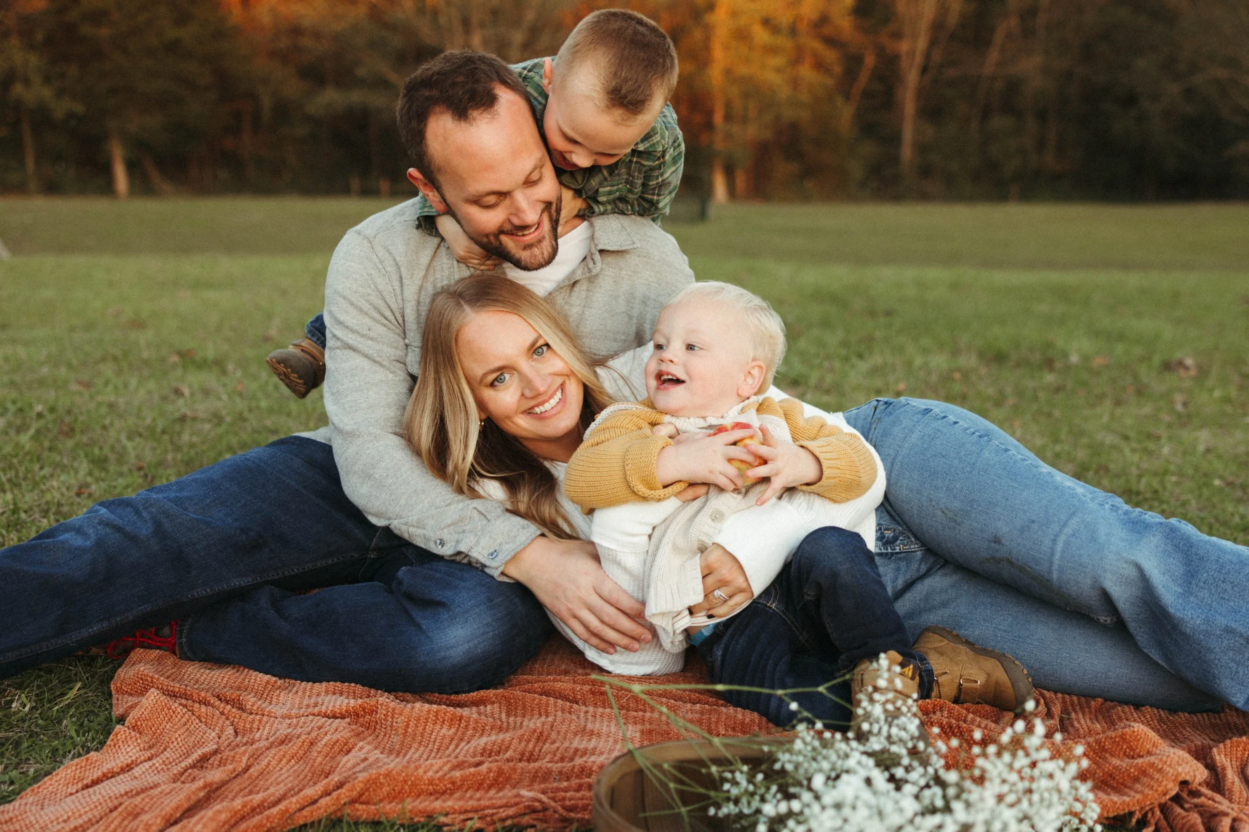 A family of four enjoying time together outdoors on a blanket in a park during autumn. The father is lying on the blanket with the mother and a young child in front, while the older son is riding on the father's back. They are smiling and playing happily.