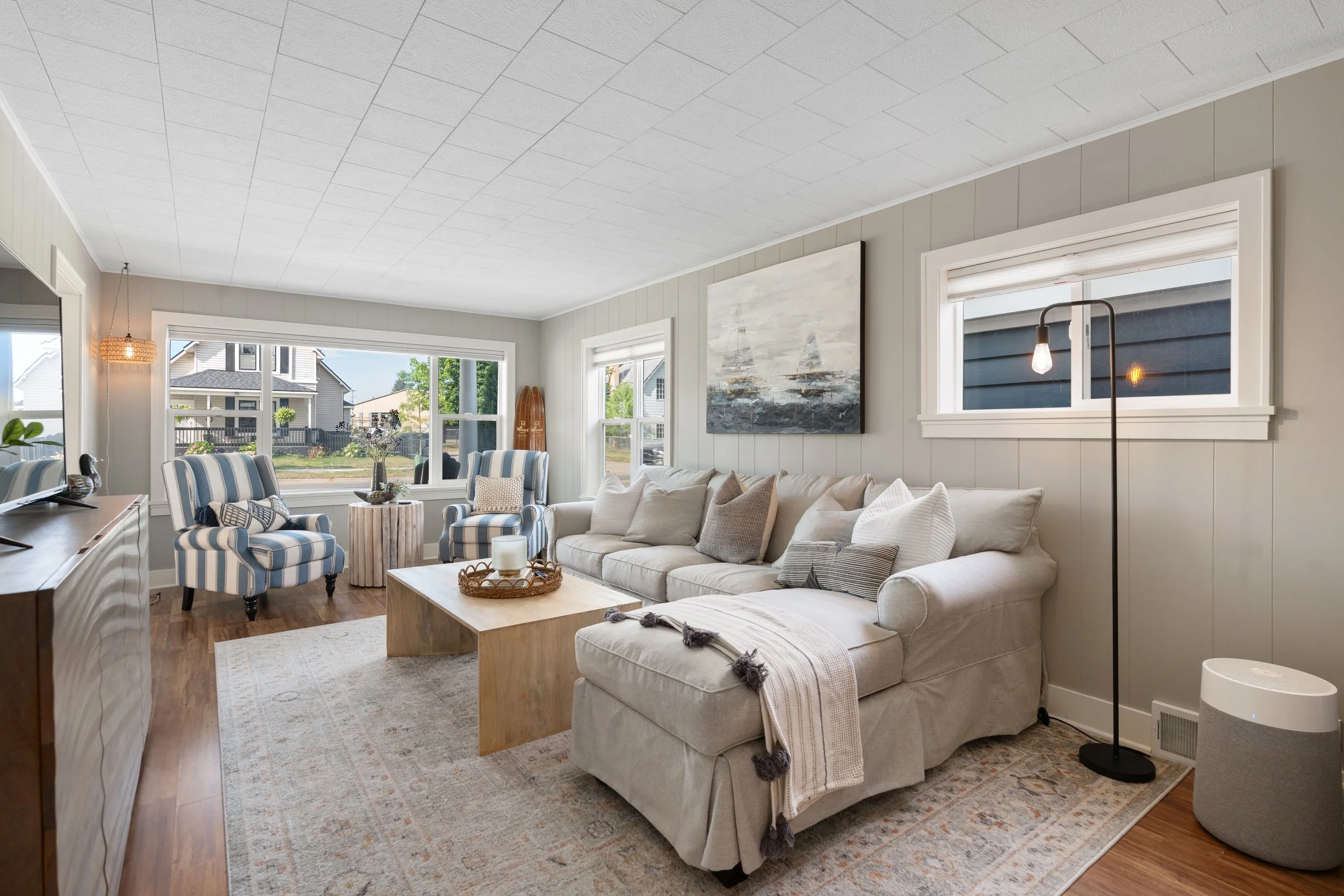 Living room with beige sofa, striped blue and white armchairs, a wooden coffee table, rugs, large windows, artwork on the wall, and a floor lamp.