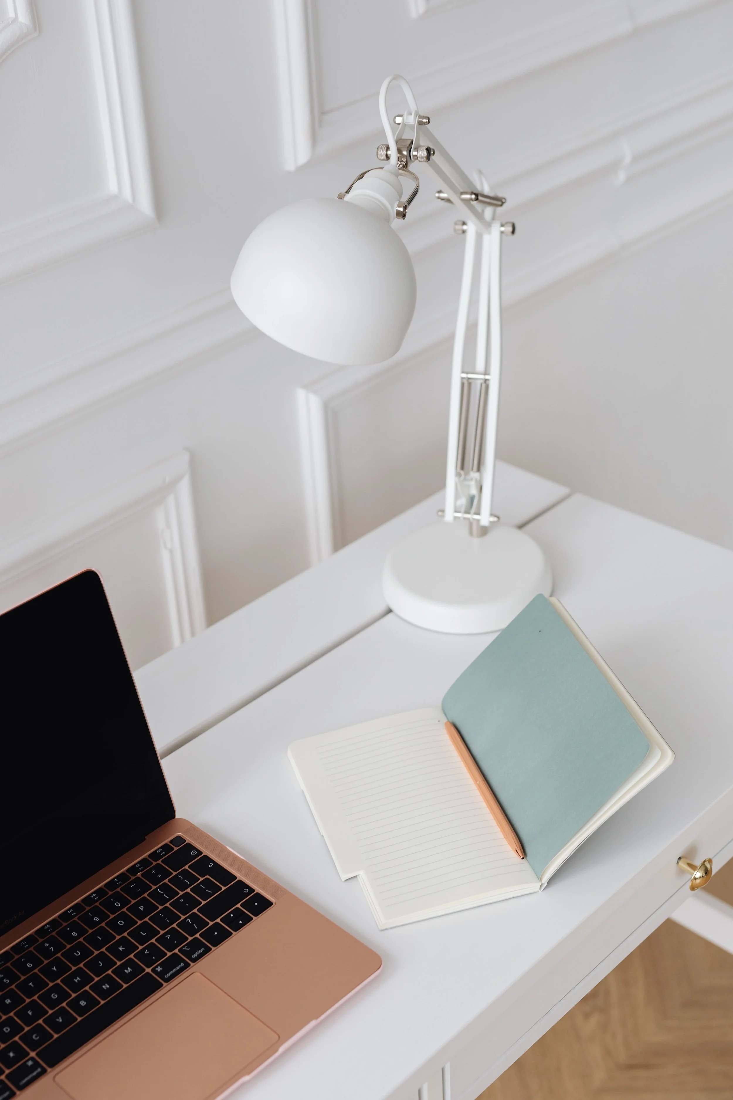 White desk with an adjustable lamp, rose gold laptop, notebook, pencil, and blue book